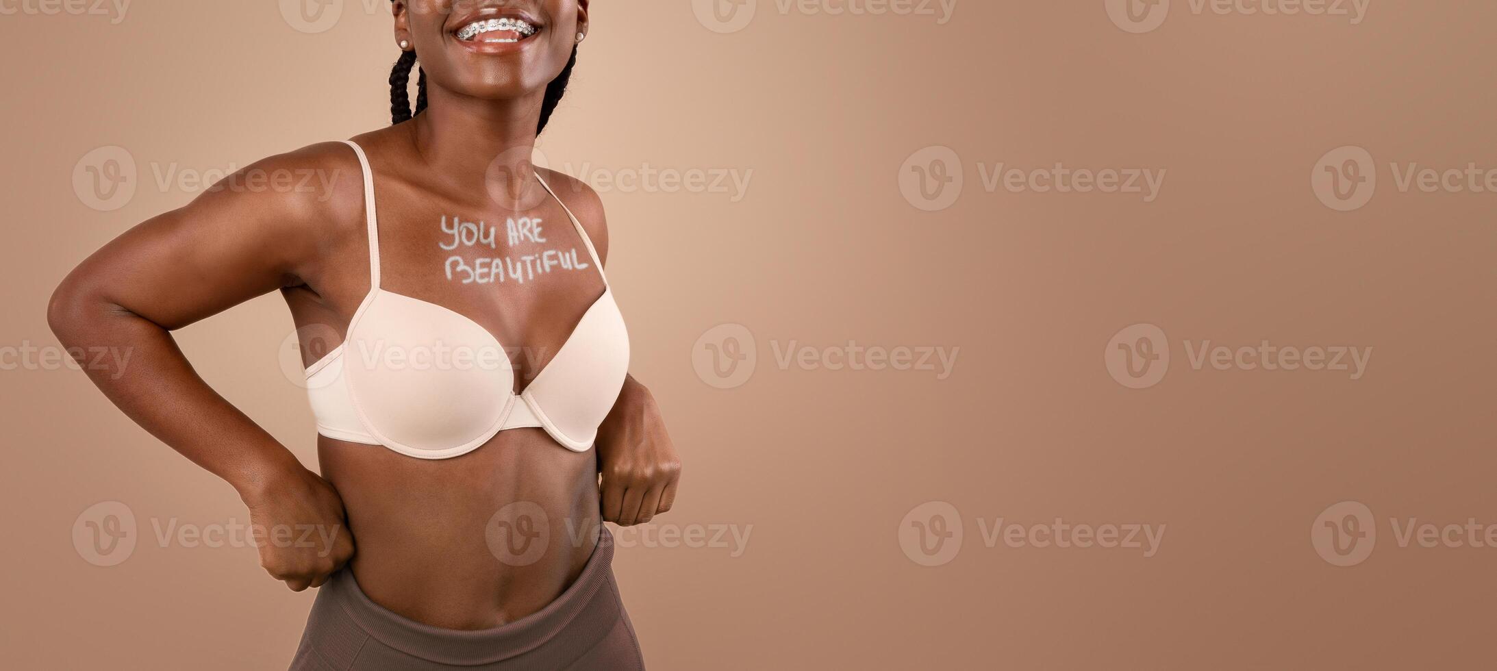 Love Yourself. Cheerful Black Slim Girl With Inscription You Are Beautiful On Chest Posing In Bra Over Beige Background In Studio, Free Space photo