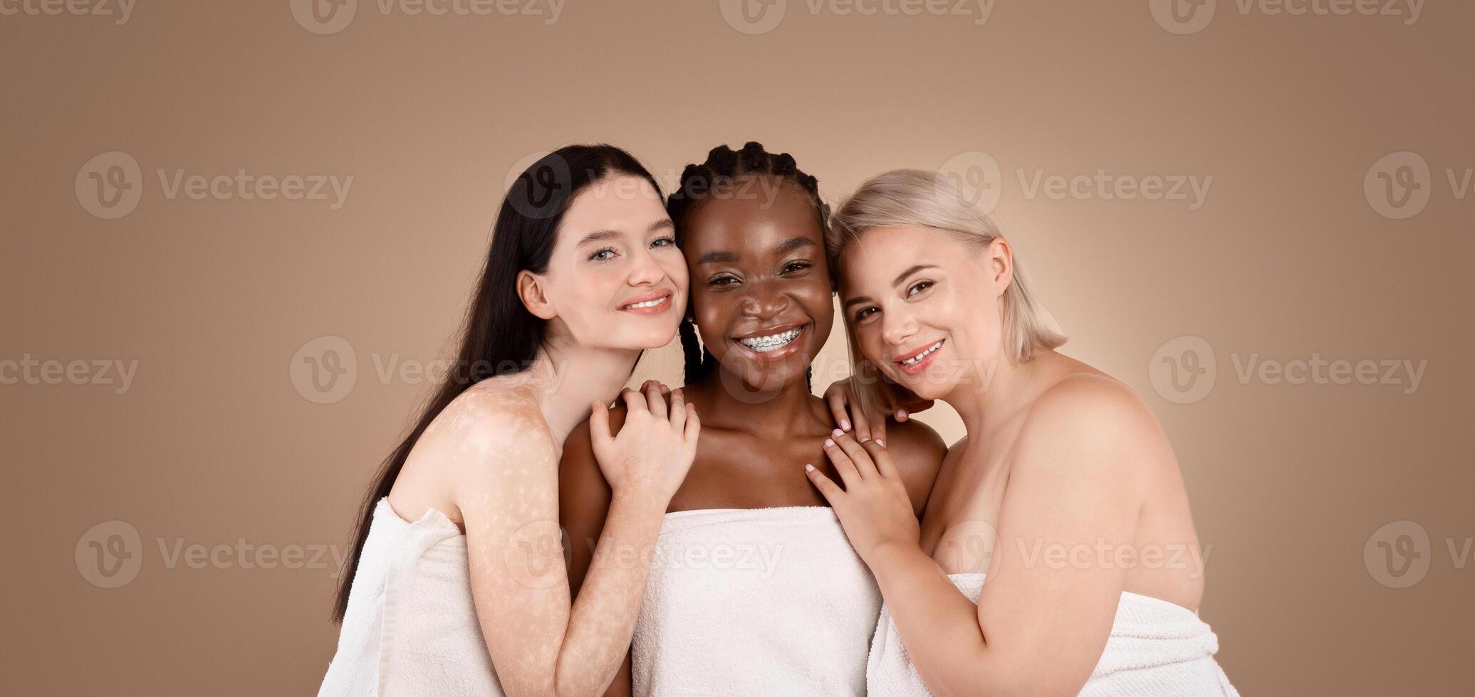 Three multiracial females with different body and skin types wrapped in towel, posing over beige studio background, smiling at camera, copy space photo