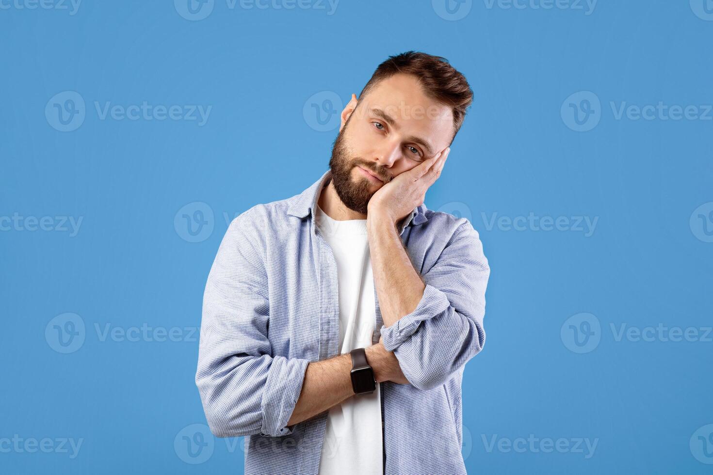 Bored young guy leaning on his hand and looking at camera over blue studio background. Tired millennial man feeling exhausted or overworked, not interested in something photo
