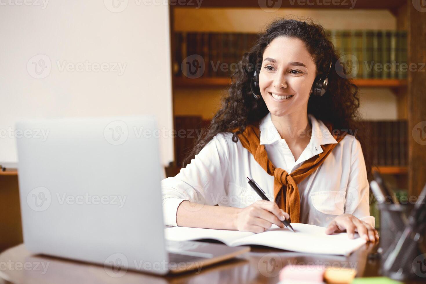 Doing Homework. Portrait of smiling young curly woman sitting at desk with pen, writing and taking notes in notebook, using and looking at laptop. Adult lady watching online course during quarantine photo