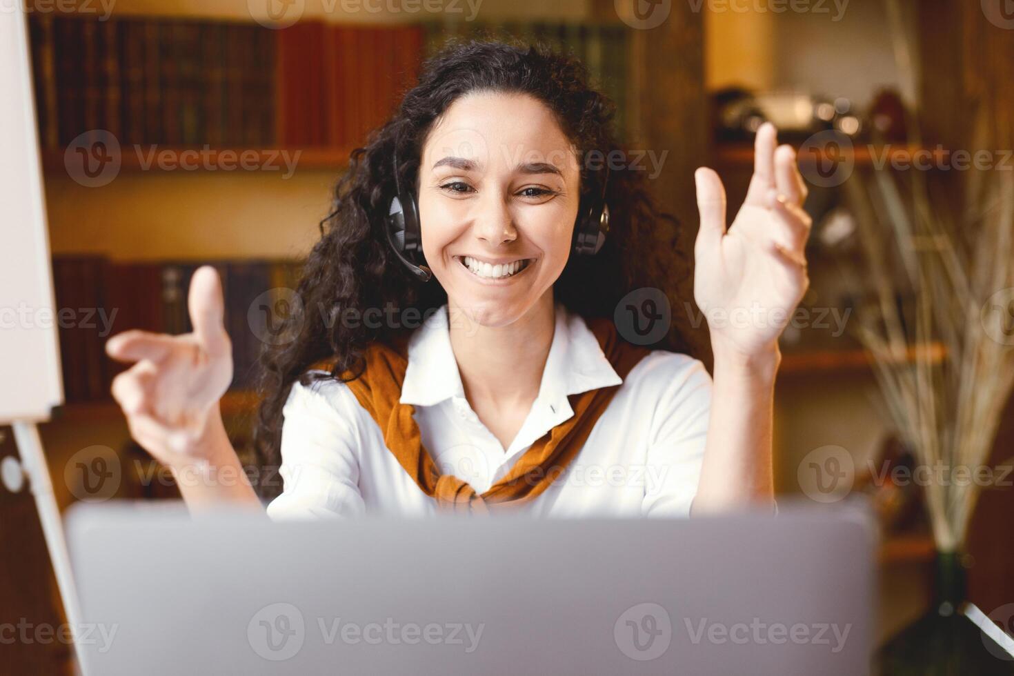 Online Call And Support Concept. Smiling female customer service representative making web conference on laptop computer, talking to camera and consulting client, sitting at table, explaining photo