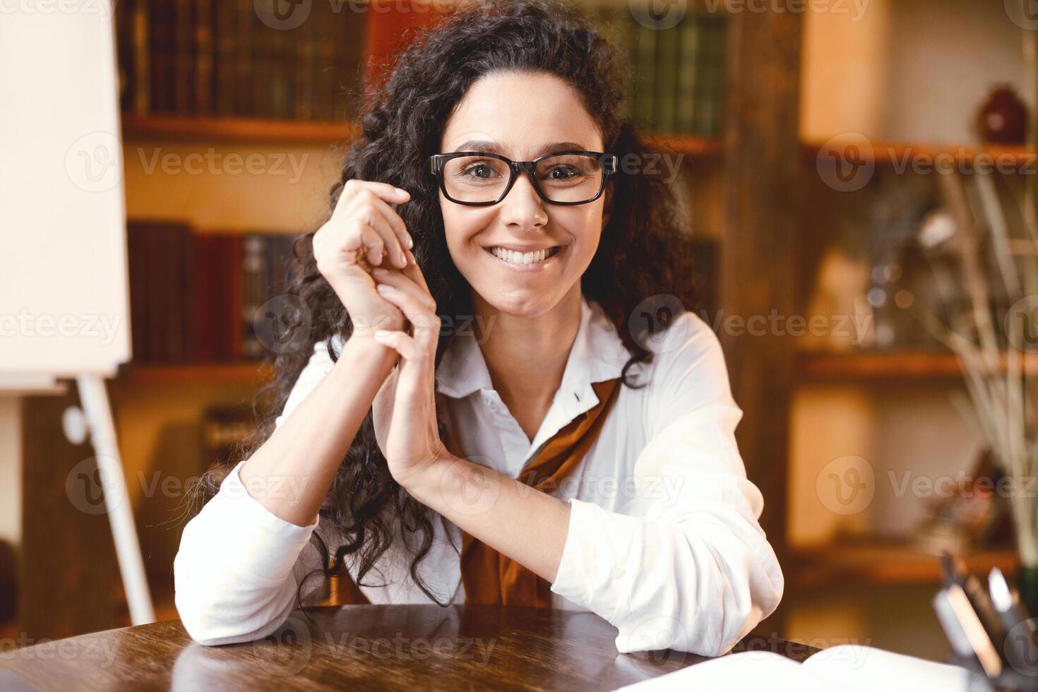 Portrait of smiling confident curly lady in eyeglasses sitting at desk with bookshelves and board in the blurred background. Casual woman in white shirt smiling and looking at camera photo