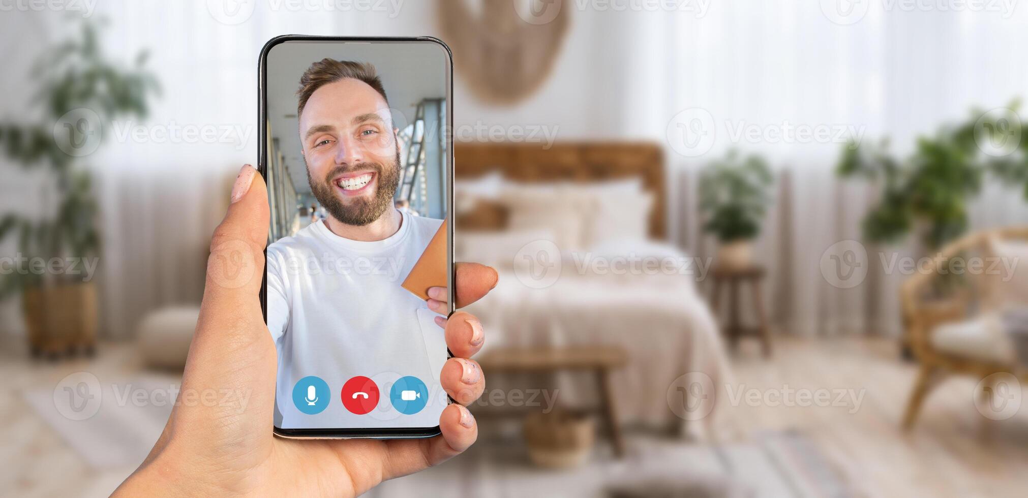 A person holding a smartphone displaying a call. The cozy home setting is decorated with plants, and natural light fills the room, creating an inviting atmosphere. photo