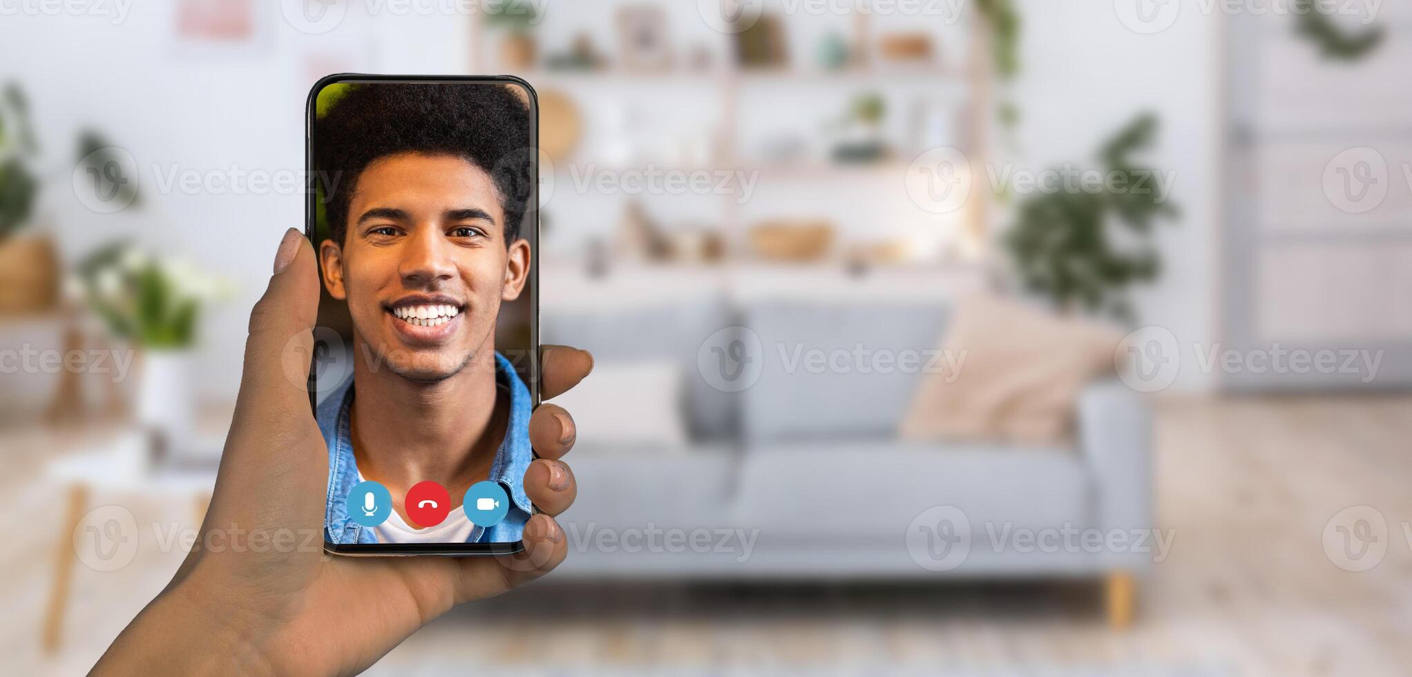 A person is holding a smartphone displaying a smiling individual during an online communication session. The cozy home setting features a stylish couch and decorative plants. photo