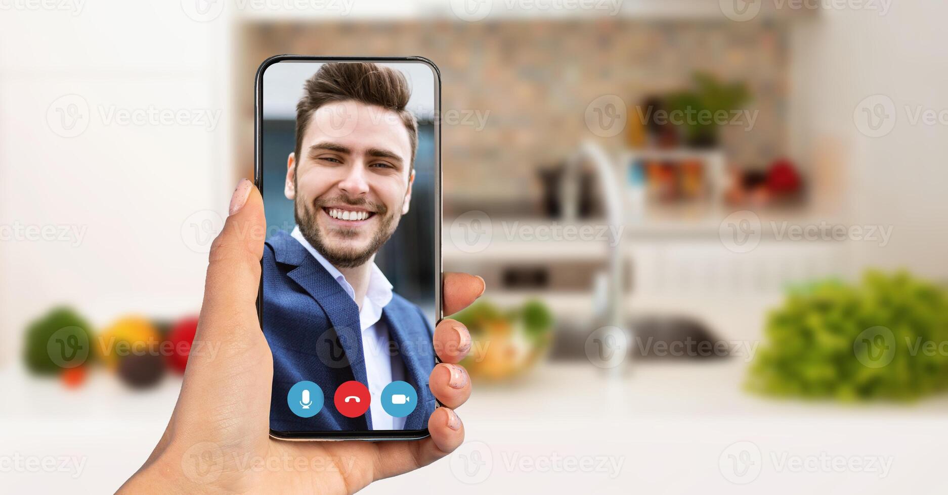A person holds a smartphone displaying a smiling friend on a call. The setting is a cozy kitchen, filled with fresh produce. The atmosphere suggests a relaxed evening chat. photo