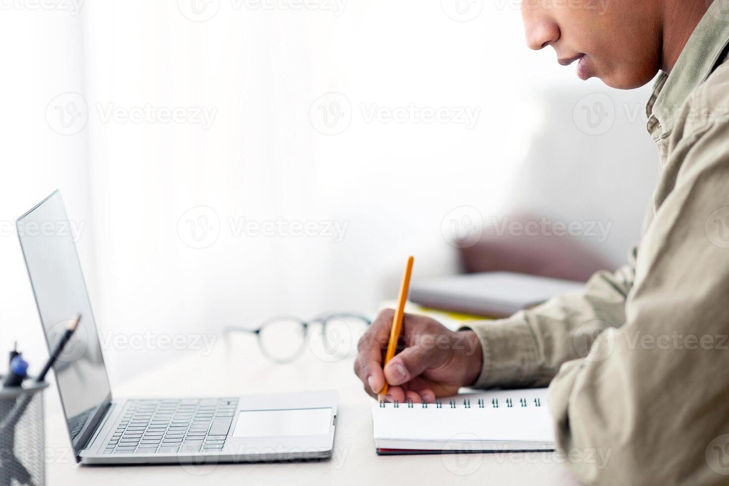 Remote education concept. Side view of black teen guy taking notes in copybook during web conference at home. African American adolescent taking online lesson or listening to lecture on internet photo