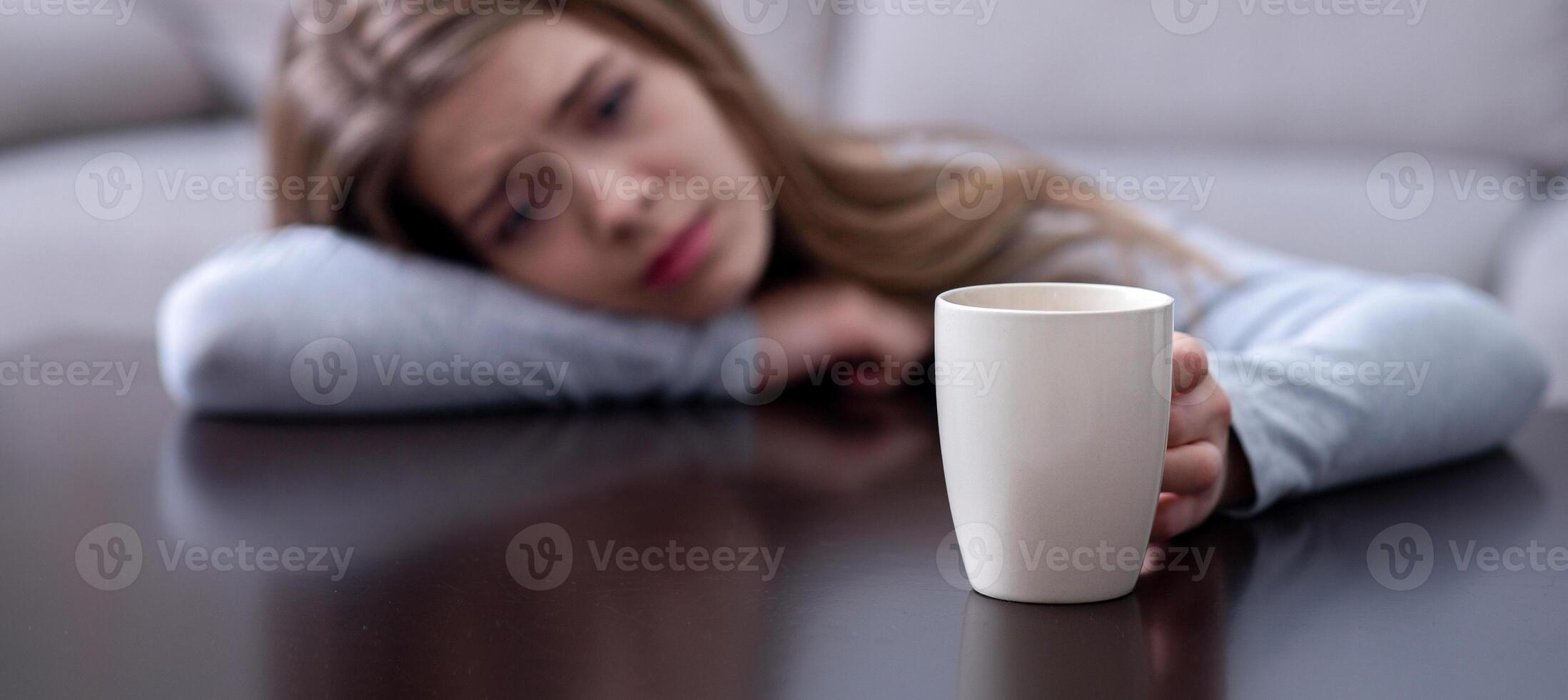 Melancholic young woman leaning on table and holding cup with hot drink, suffering from depression. Selective focus. Copy space. Mood swings or psychological problems concept photo