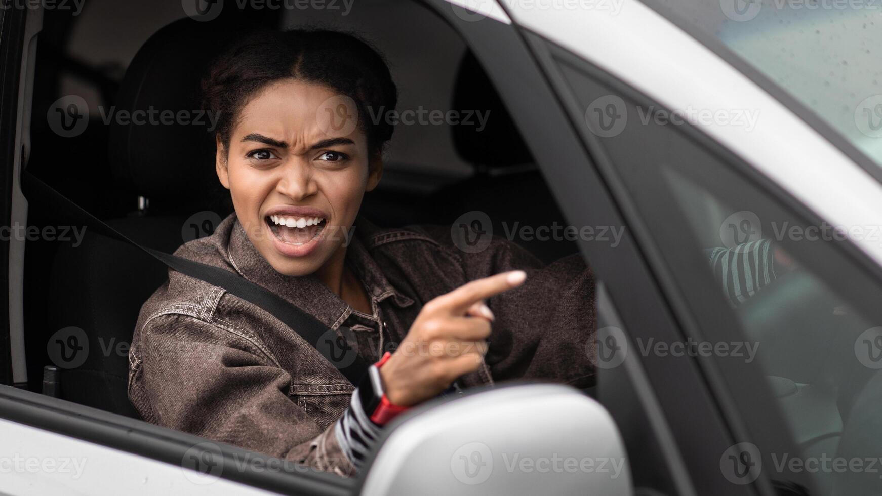 Angry, young, pissed off, displeased, aggressive african american woman shouting on drivers in front of her and points finger, driving car. Road traffic jam, negative human emotions and scandal photo