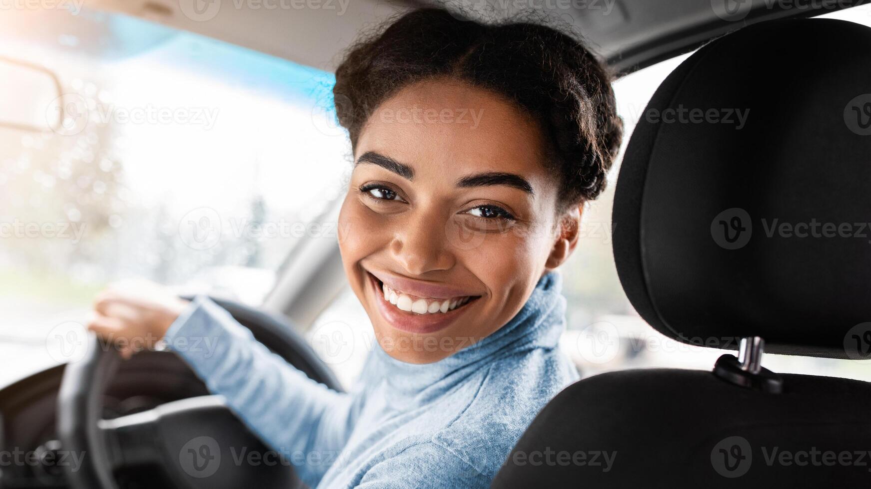 Driver turns to passenger seat and smiles at camera. Happy millennial african american woman taxi driver sitting in car holding to steering wheel, next to smartphone with blank screen, panorama photo