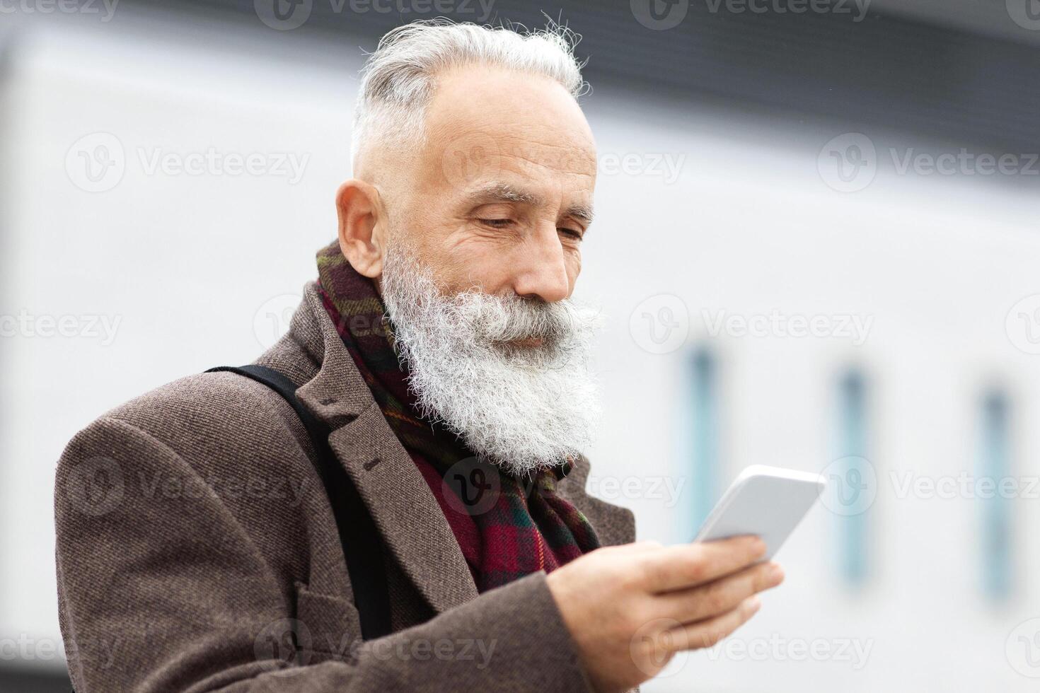 Senior grey-haired businessman using smartphone on the street, checking his agenda, copy space. Bearded aged man using mobile phone, websurfing or chatting with colleagues, having lunch break photo