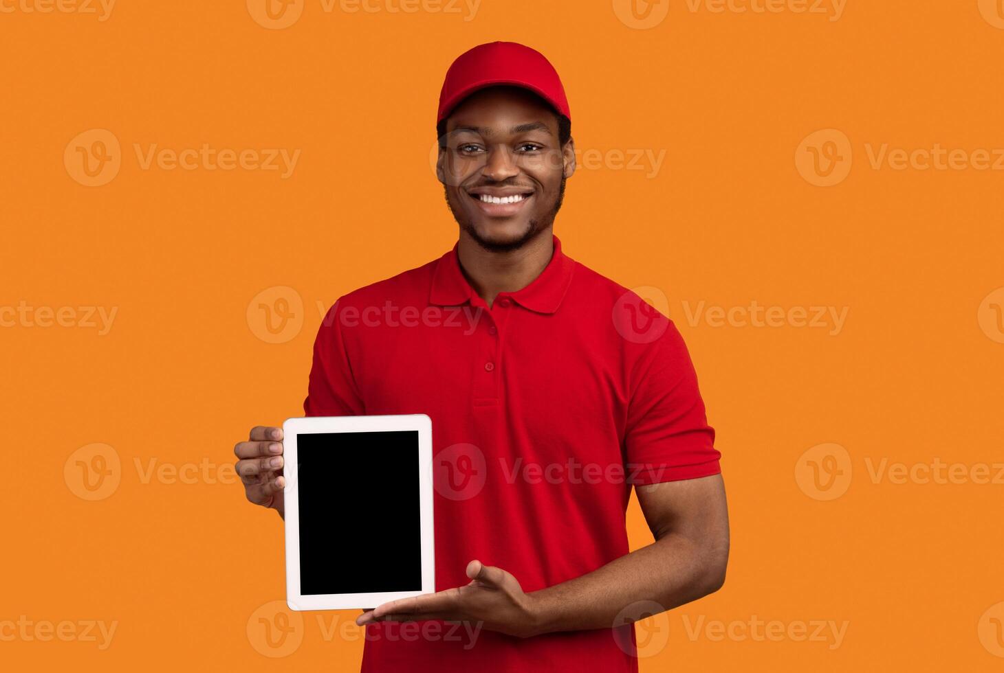 Delivery Concept. Portrait of smiling black male courier holding digital tablet with empty screen for mock up and showing to camera, wearing red cap and uniform, standing isolated on orange background photo