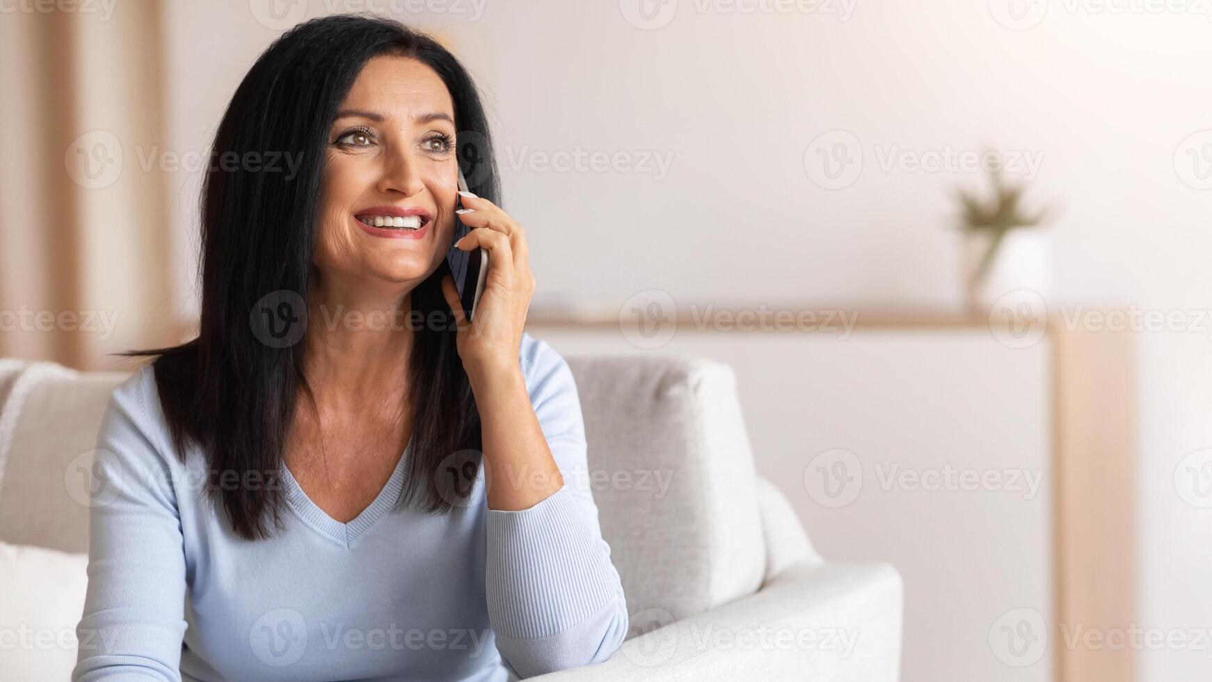 Phone Call. Portrait of smiling mature lady talking on cellphone, resting on couch in living room at home. Communication Concept. Cheerful woman having pleasant conversation with her children photo