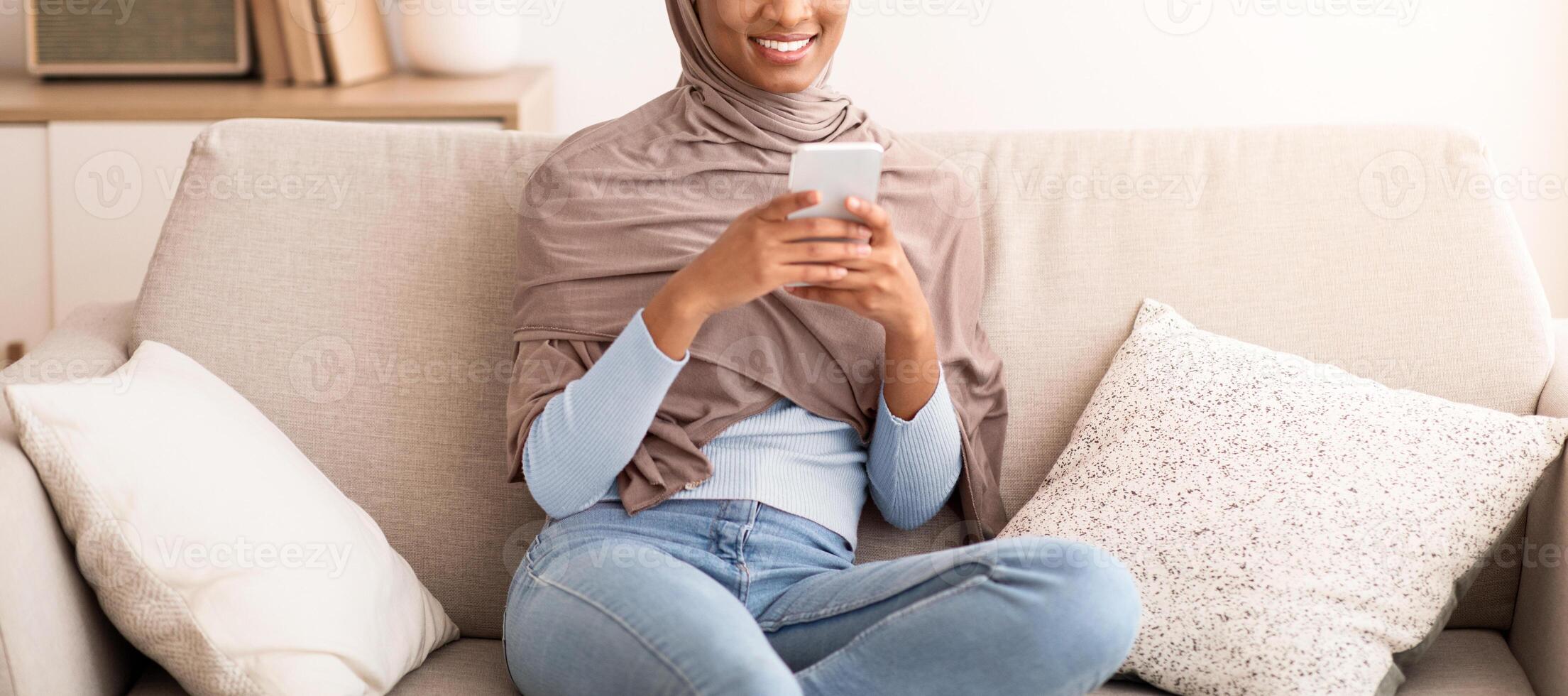 Pretty Arab woman in hijab texting on smartphone, checking social media, sitting on sofa at home. Lovely black lady in Muslim headscarf using cellphone for work or entertainment photo