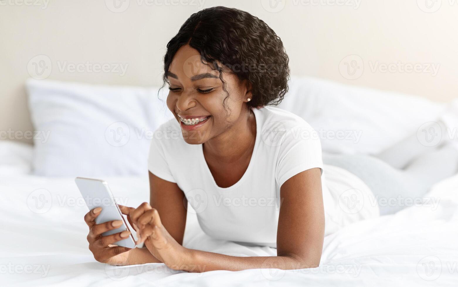 Joyful black lady laying on bed at home, using mobile phone and smiling, copy space. African american woman spending weekend at home, chatting with friends or checking social media on smartphone photo