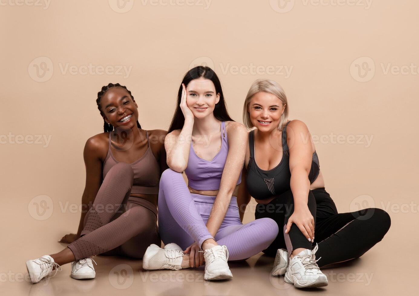 Portraif Of Three Different Multi Ethnic Ladies Posing In Sportswear, Happy Body Positive Females With Different Sized And Appearances Sitting On Floor On Beige Background, Smiling At Camera photo