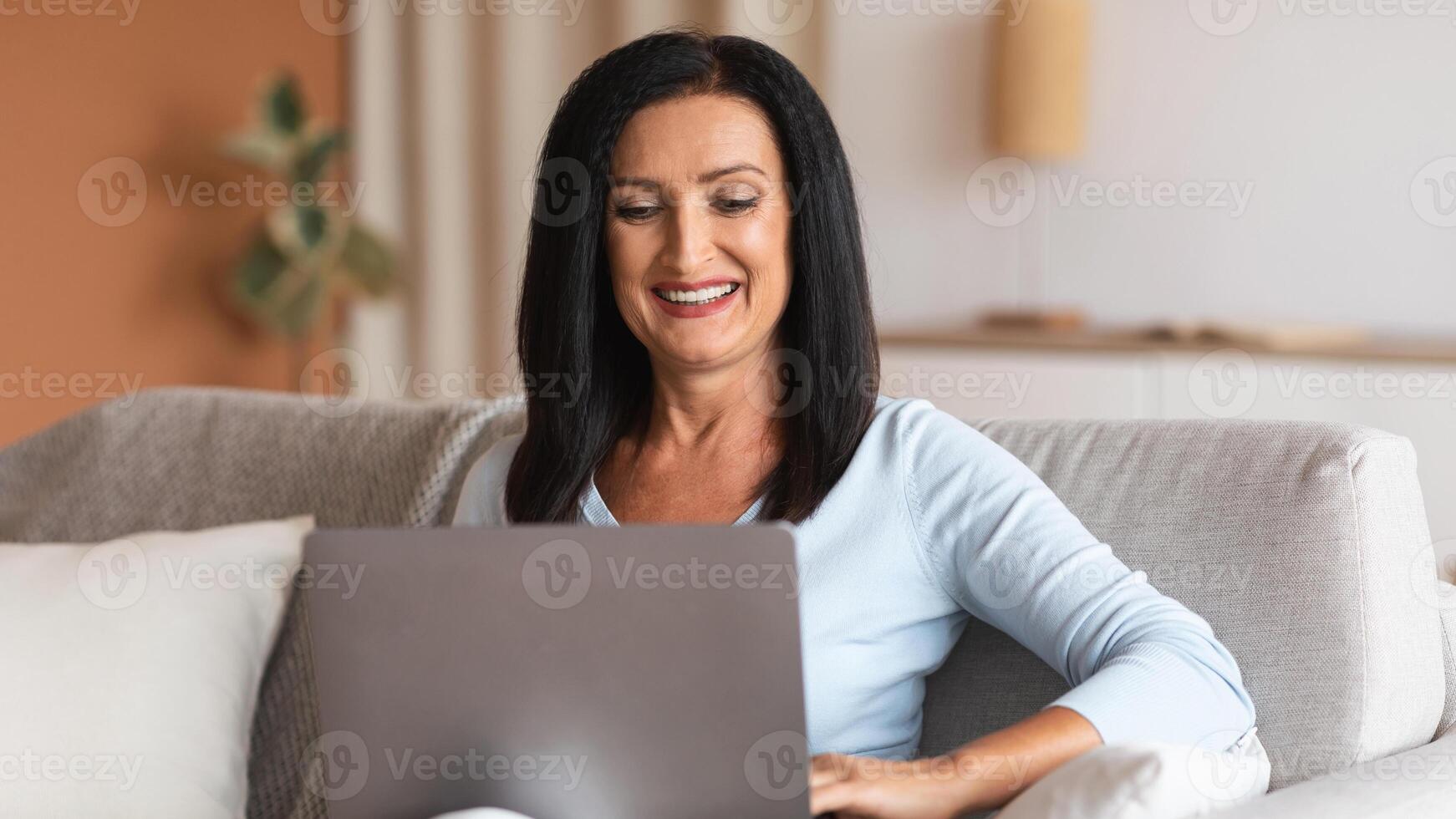 Domestic Comfort. Portrait Of Positive Mature Woman Using And Working On Laptop At Home, Enjoying Free Time, Smiling Lady Sitting On The Couch With Computer On Her Lap, Watching photo
