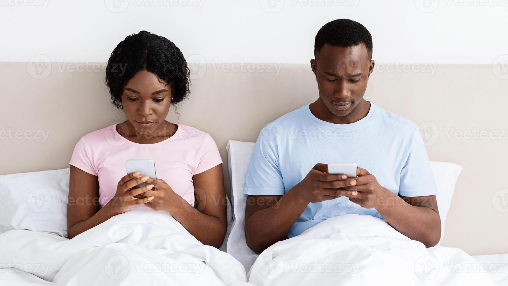 Black young man and woman sitting in bed and using their smartphones before sleeping, indifference in relationships concept. African american married couple experiencing crisis in their marriage photo