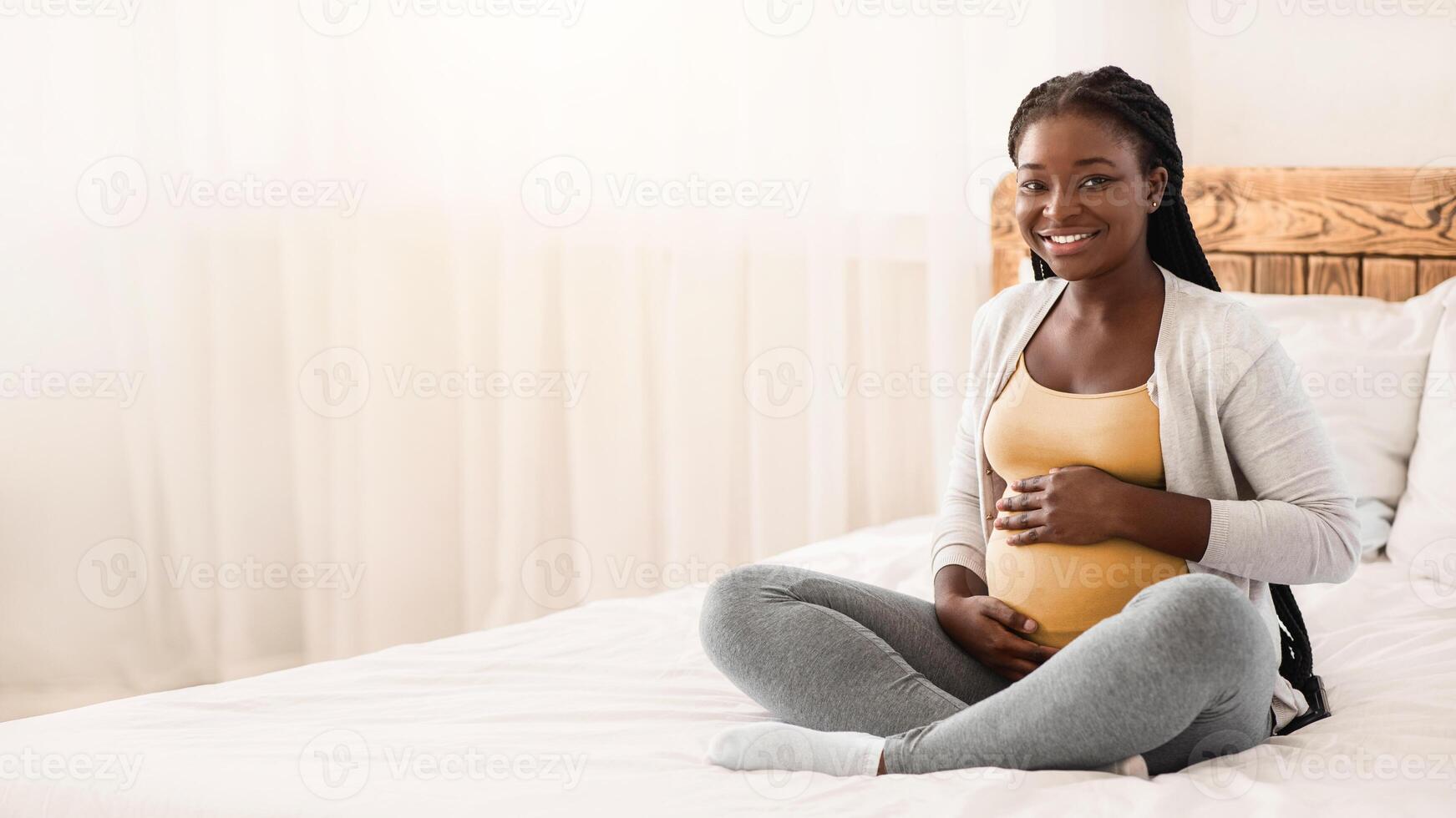 Pregnancy Time. Happy Black Pregnant Woman Sitting On Bed Touching Belly, Young Expectant Lady Posing In White Bedroom, Cuddling Tummy And Smiling At Camera, Resting At Home, Copy Space photo