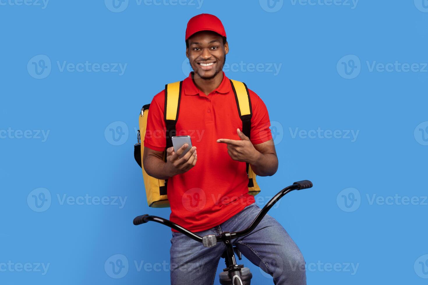Courier Service Application. Smiling black delivery guy using smartphone and pointing at it, wearing red uniform, cap and thermo bag, riding bicycle, isolated on blue studio background photo