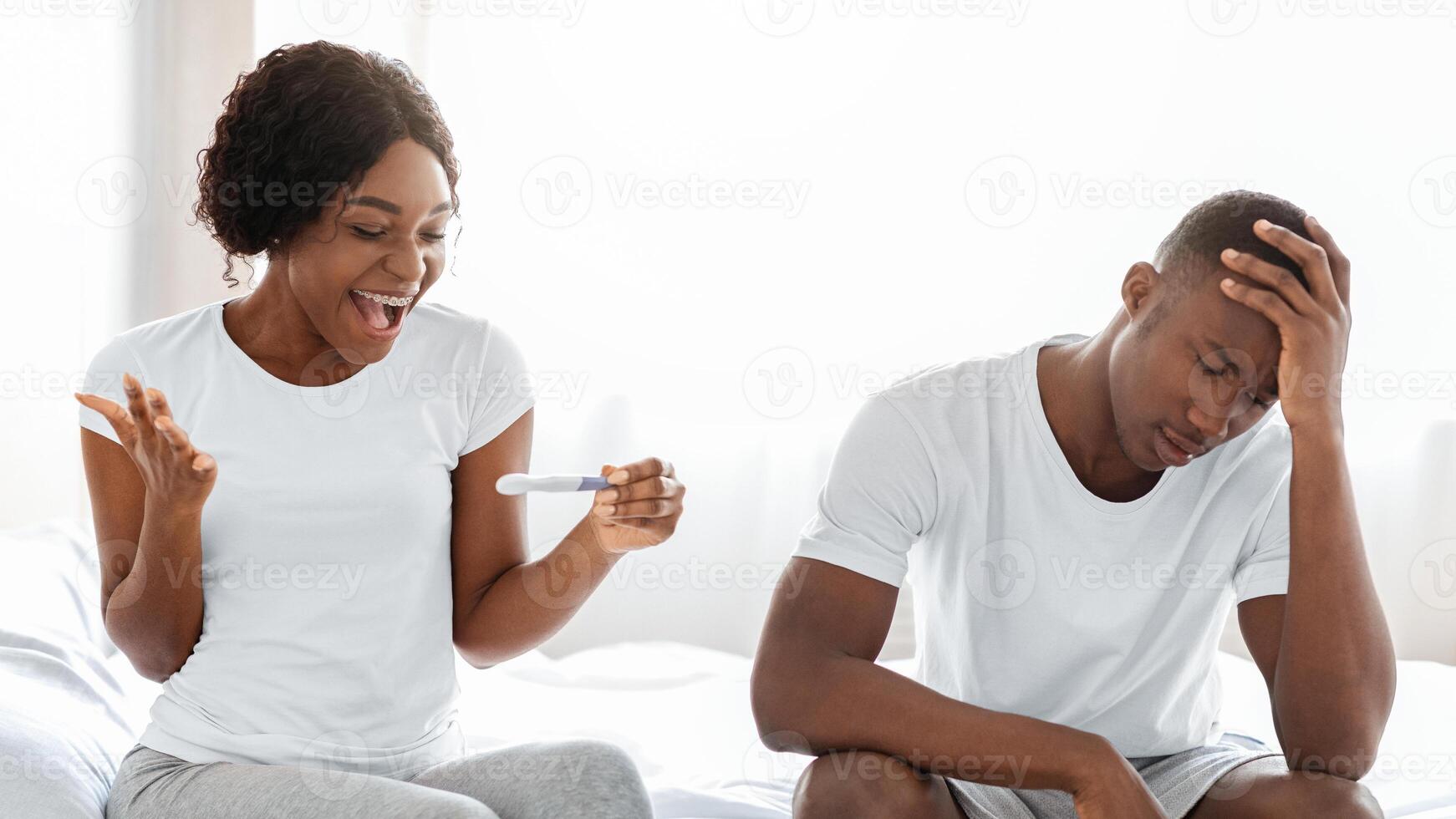 Happy black woman with pregnancy test celebrating positive result and her shocked confused boyfriend touching his head, african american couple sitting on bed at home, copy space photo