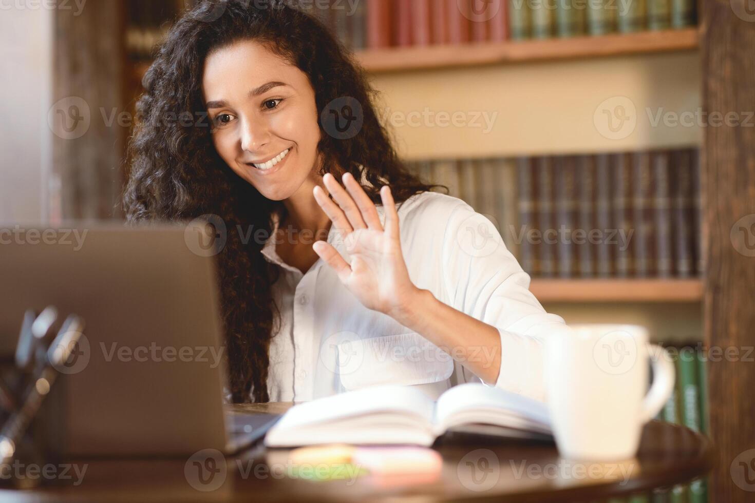Virtual Conference. Portrait of smiling young woman making online call on laptop, waving with hand to webcam. Communication Concept. Woman chatting using computer, sitting at desk photo