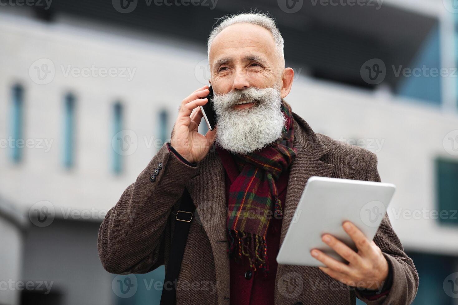 Handsome grey-haired man talking on mobile phone while going by street, holding digital tablet, copy space. Good looking elderly businessman having business conversation on phone photo