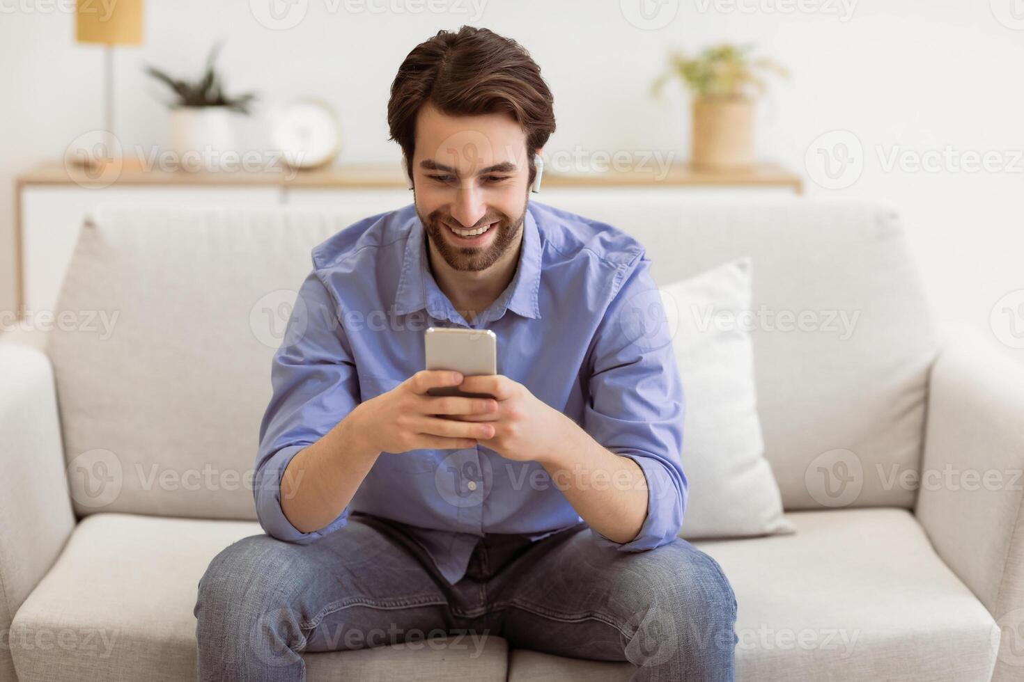 Happy Man In Earbuds Making Call On Smartphone Sitting On Sofa At Home. Guy Texting Using Phone In Living Room Indoor. Technology And Gadgets, Great Mobile Application Concept photo