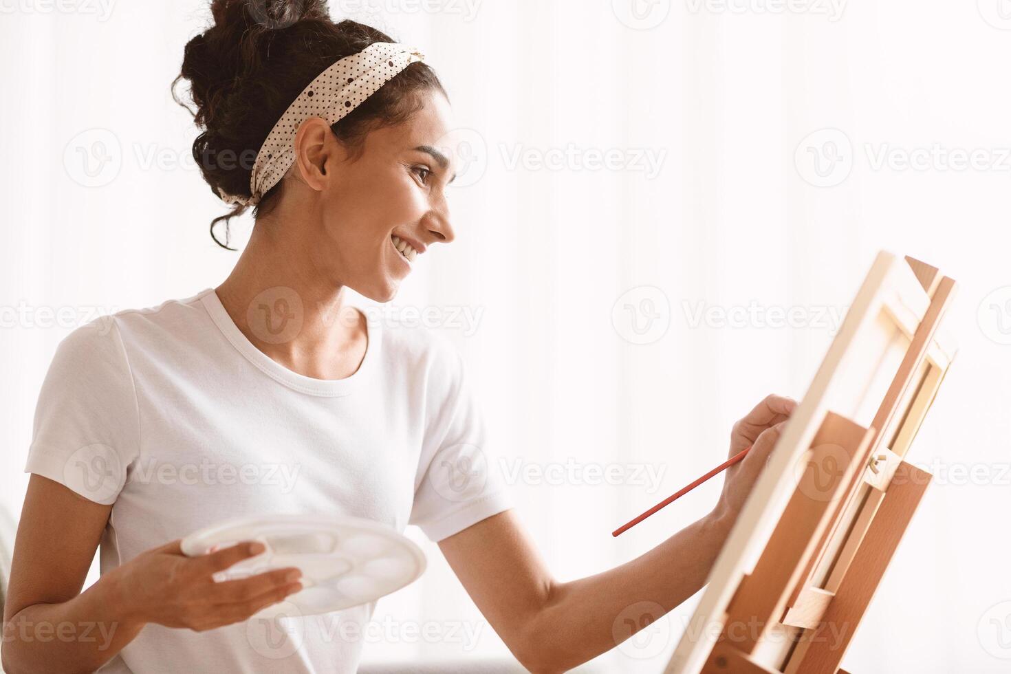 Young smiling woman artist painting at home, using easel, palette and paintbrush, side view, copy space. Closeup of creative lady drawing while having free time during quarantine photo