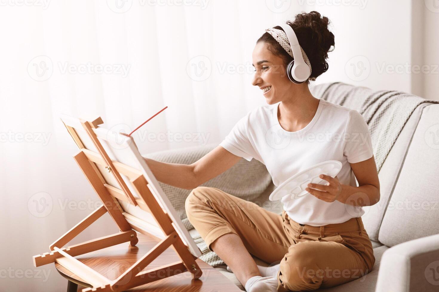Joyful youg woman with wireless headphones listening to music while drawing at home, holding brush and palette, sitting on sofa in front of small easel, spending her free time alone, enjoying painting photo
