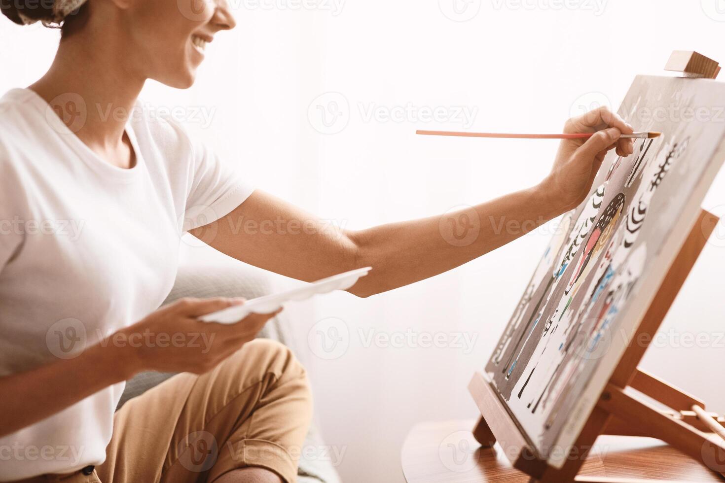 Cropped of creative lady painting at home, using easel, brush and palette, side view. Unrecognizable woman enjoying her free day, sitting on couch in living room and drawing, closeup. Hobby concept photo