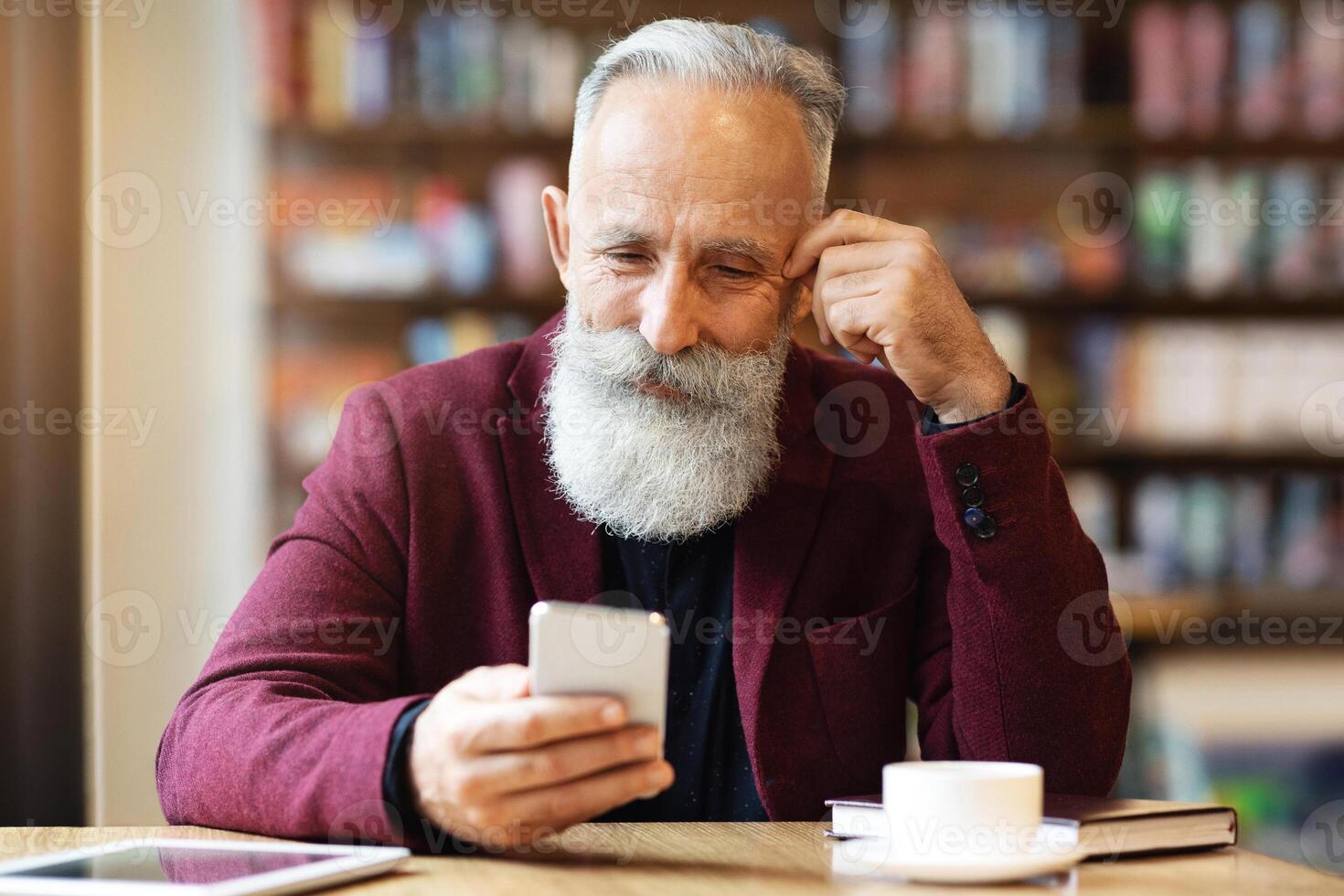 Senior grey-haired man with long beard drinking coffee at cafe, using smartphone, planning his day, copy space. Handsome aged businessman having coffee break, holding mobile phone photo