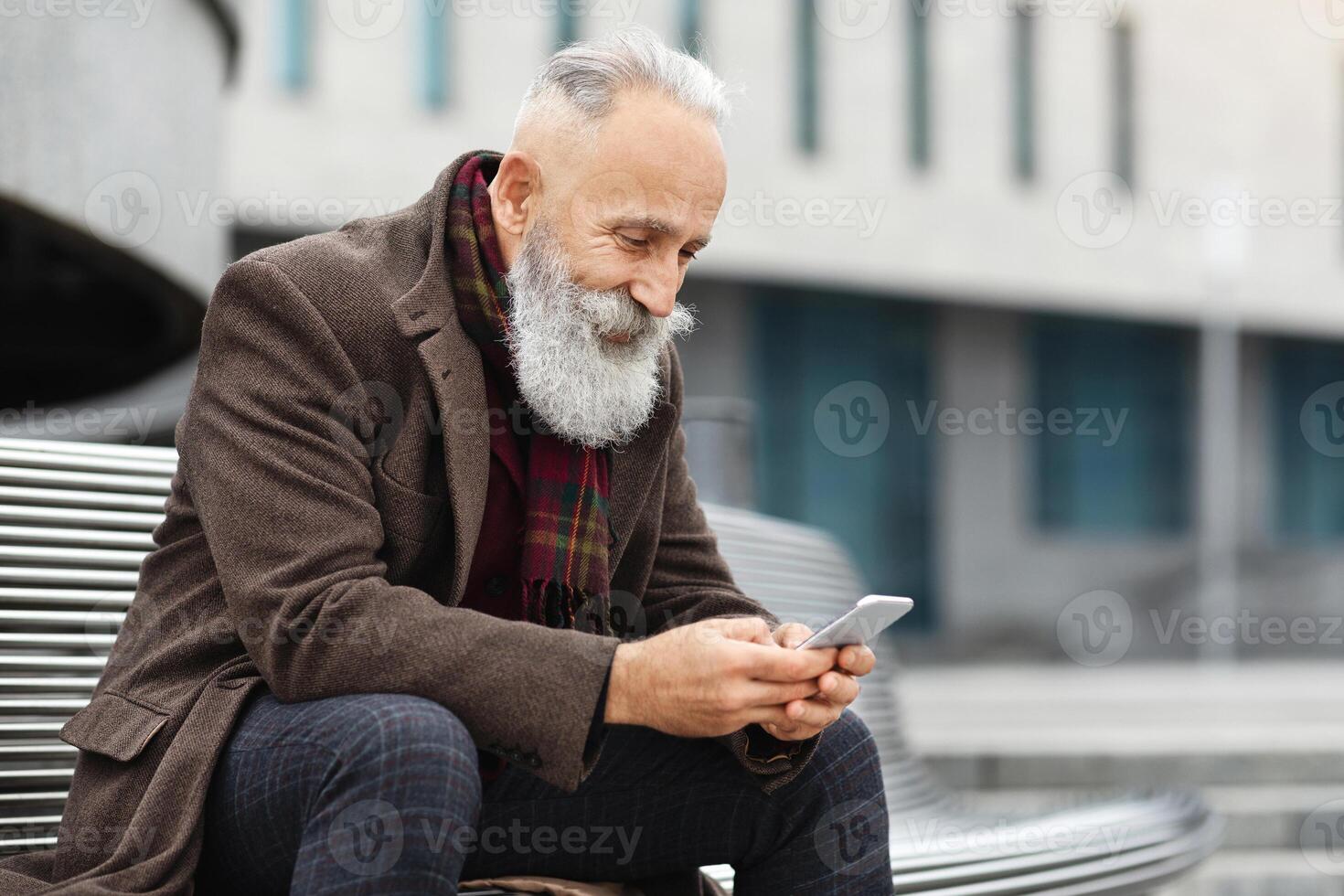 Happy elderly grey-haired man using smartphone while resting on street, sitting on bench and using mobile application, copy space. Handsome senior man using smartphone outdoors photo