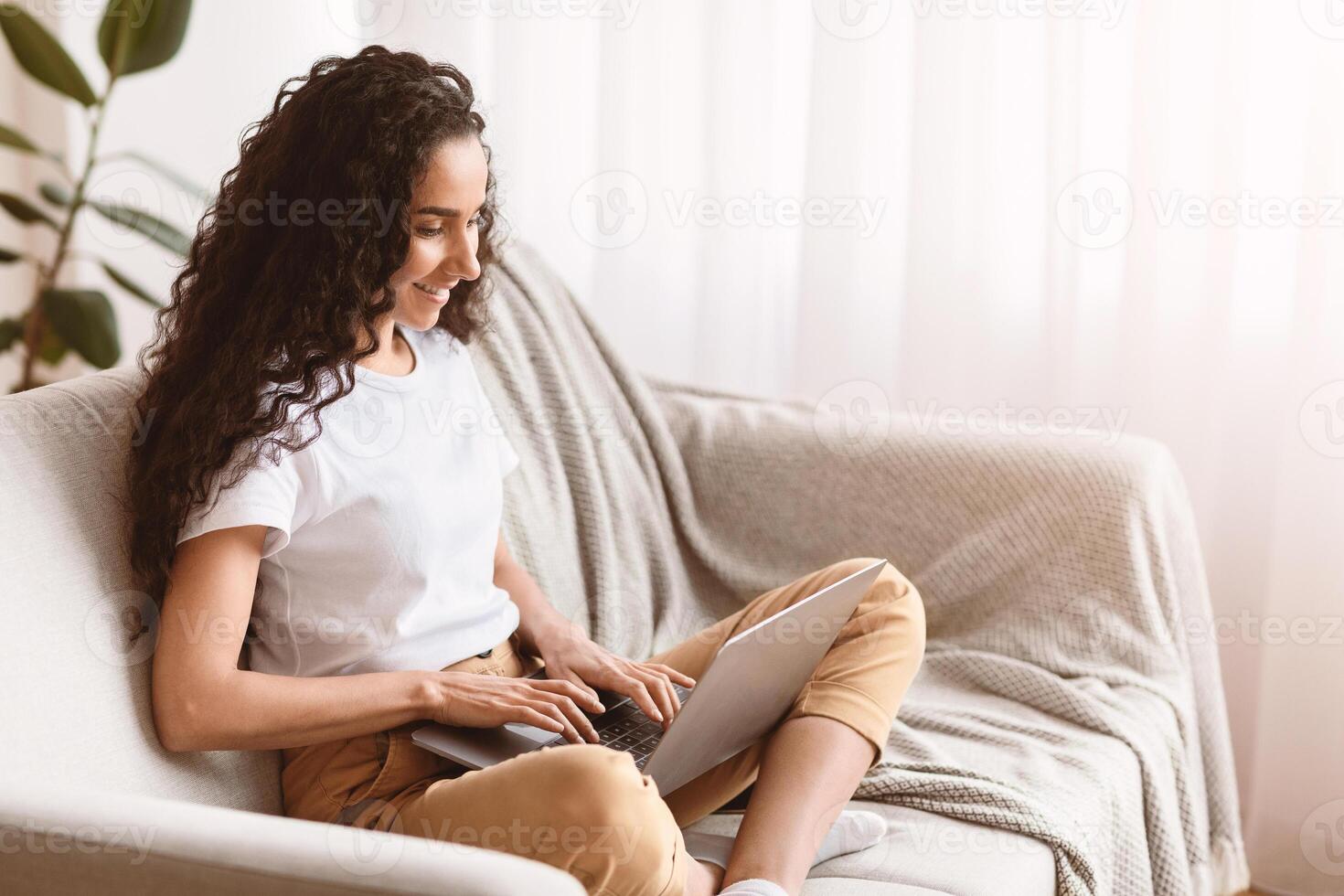 Young woman with dark curly hair freelancer working from home, sitting on couch and using laptop, typing text or communicating with clients online, blogging or copywriting, copy space photo