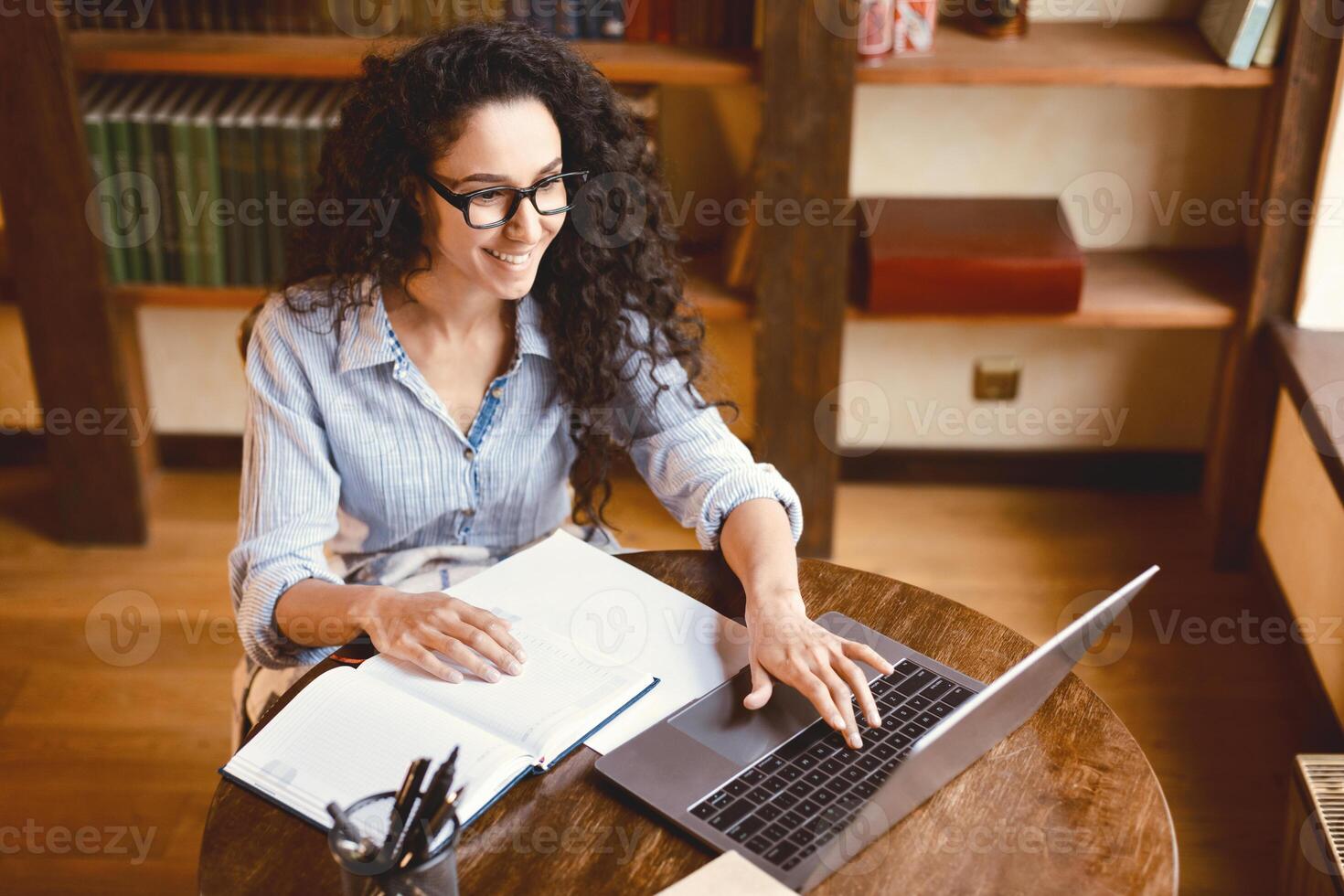 High angle view portrait of smiling young woman sitting at desk, typing on keyboard and using laptop computer. Lady watching online tutorial, webinar or seminar, studying at home, working remotely photo