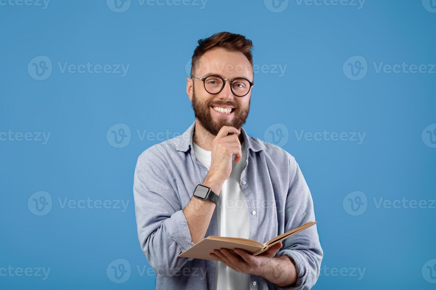 Happy young man with book looking at camera and smiling on blue studio background. Positive millennial student in casual clothes reading textbook, getting ready for college exam photo