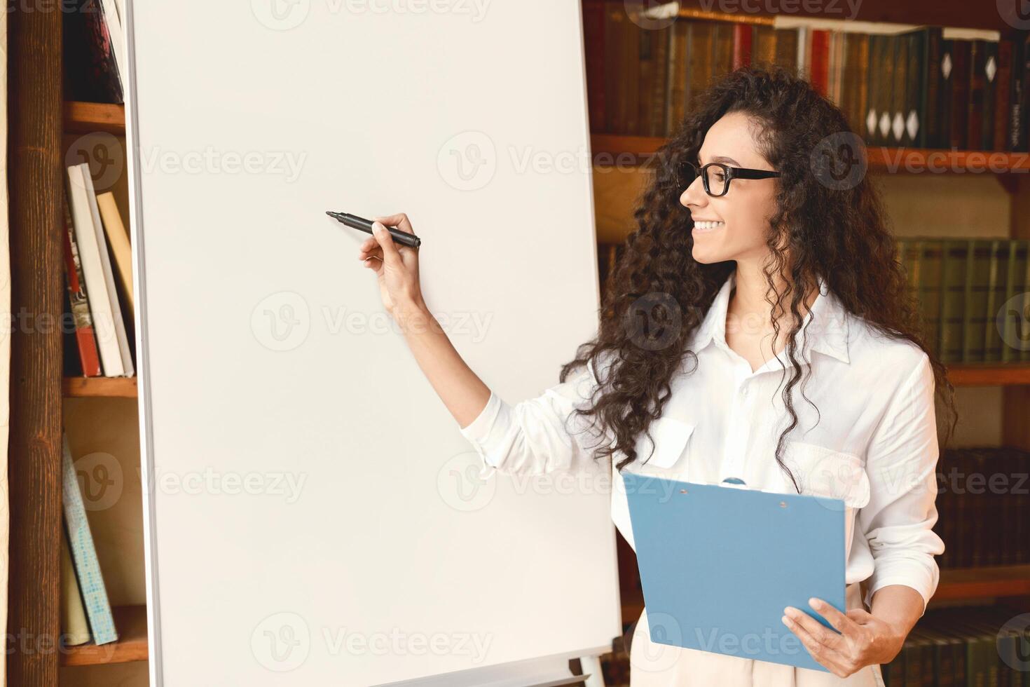 Presentation Concept. Portrait of smiling professional female manager in eyeglasses posing near blank empty whiteboard. Woman in white shirt standing with marker and writing on flipchart, copy space photo