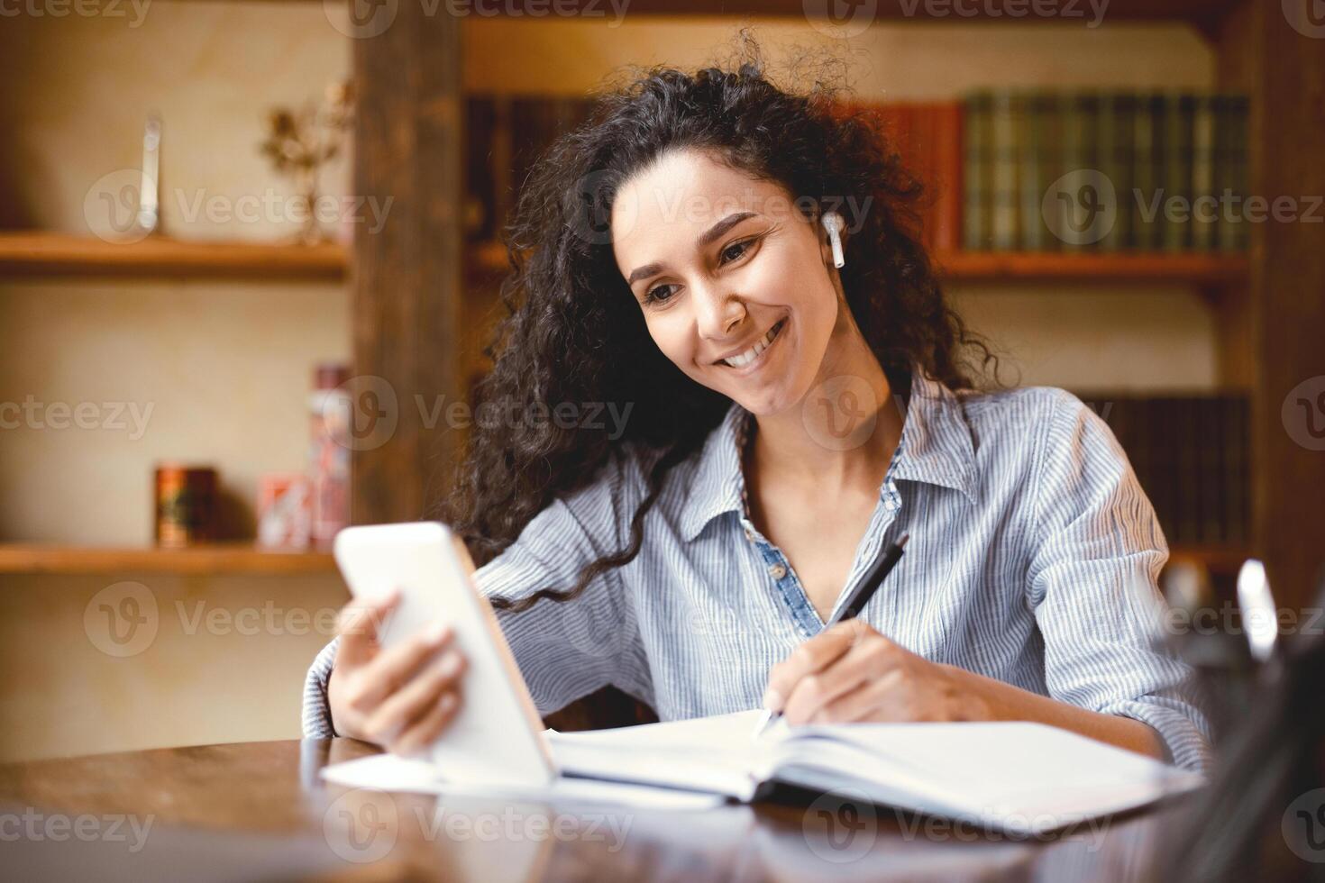 Remote Education. Smiling female student in wireless earphones sitting at desk, using smartphone and writing in notebook, taking notes, watching lesson, lecture or webinar, studying online at home photo