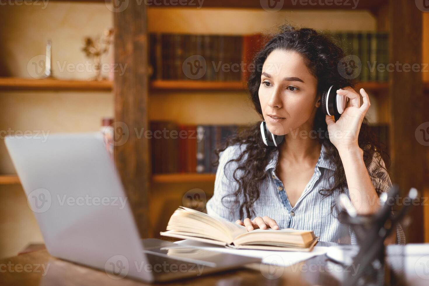 Distance Education. Portrait of woman touching wireless headset sitting at desk, using laptop and reading book. Lady watching tutorial, lecture or webinar, studying online at home. E-learning photo