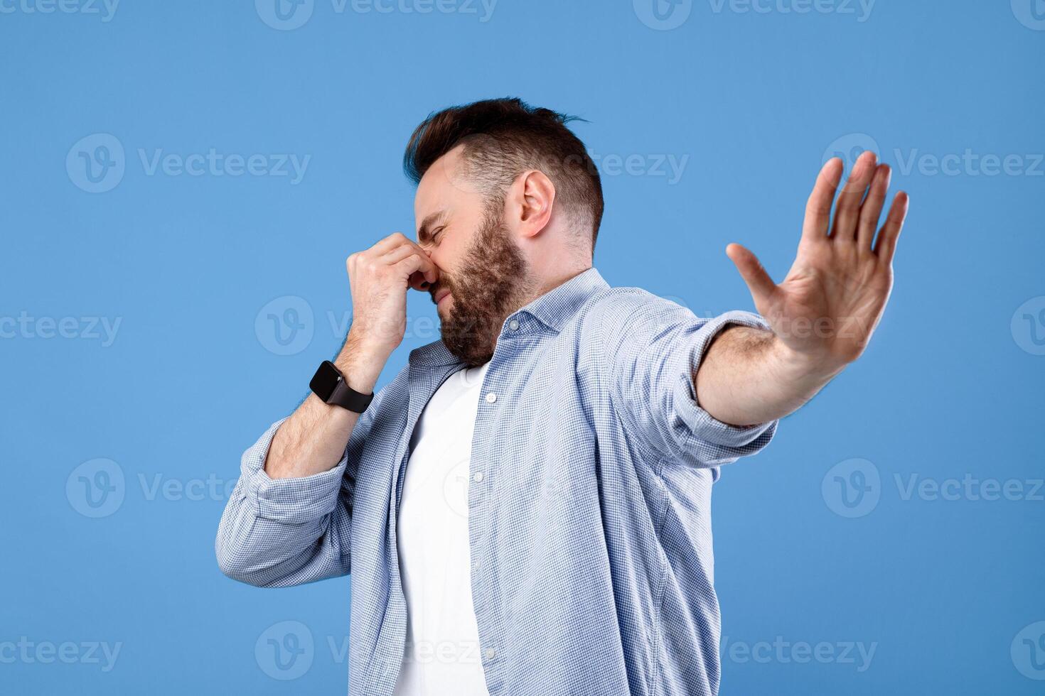 Bad smell concept. Young bearded guy closing his nose and showing STOP gesture, expressing disgust on blue studio background. Millennial man repulsed by toxic odor or terrible perfume photo