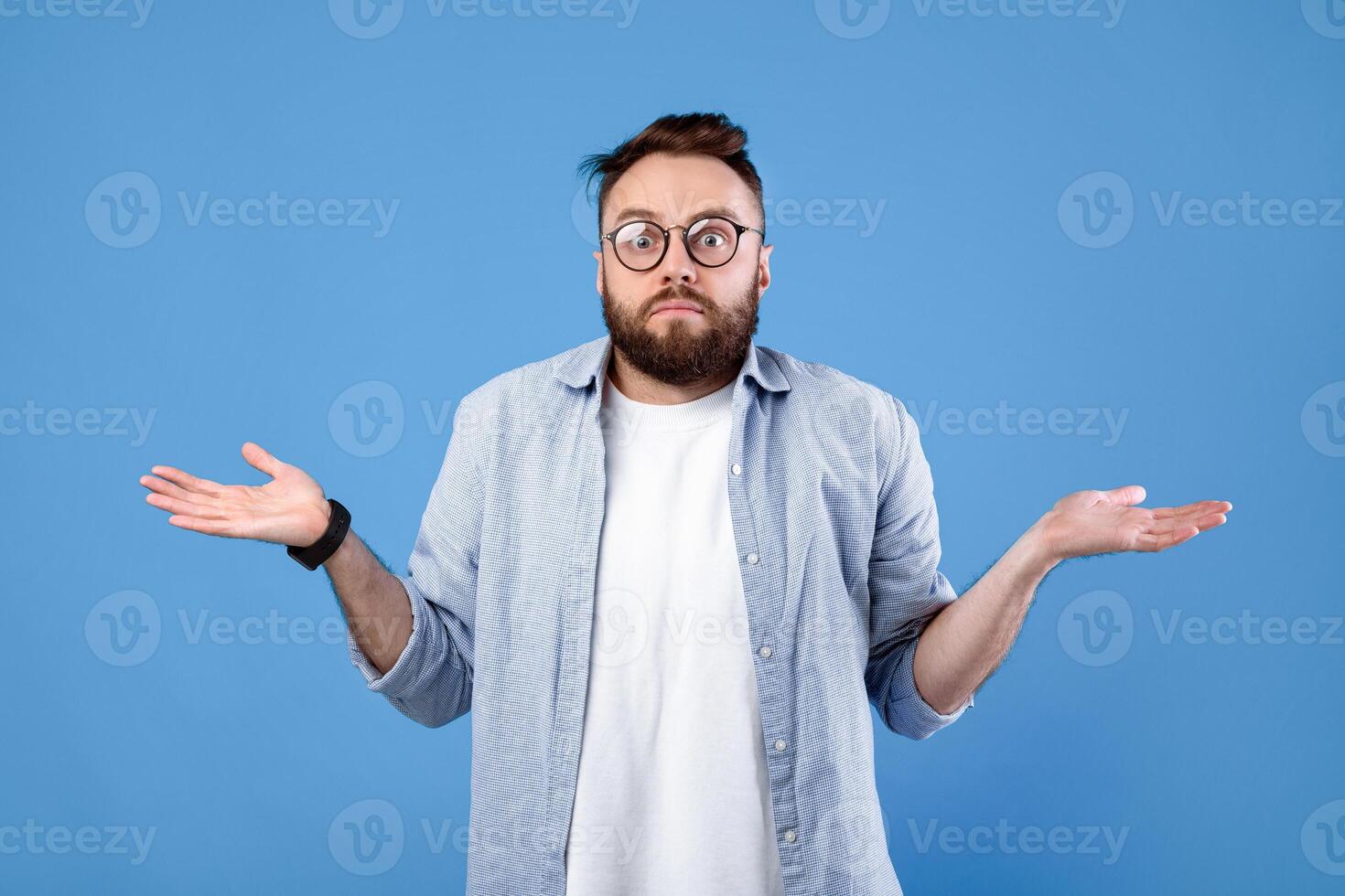 Who knows. Confused bearded man in glasses shrugging shoulders on blue studio background. Unsure millennial guy expressing uncertainty, feeling clueless, making hard choice photo