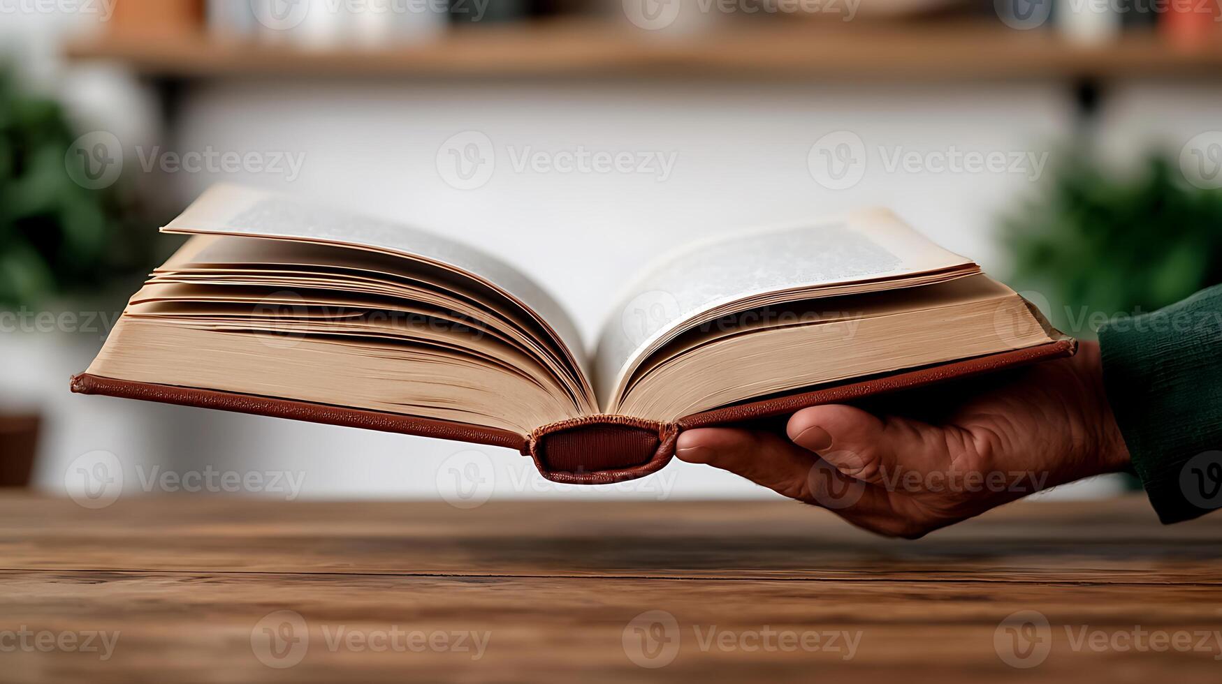 A hand holding an open book over a wooden table with blurred plants in the background photo