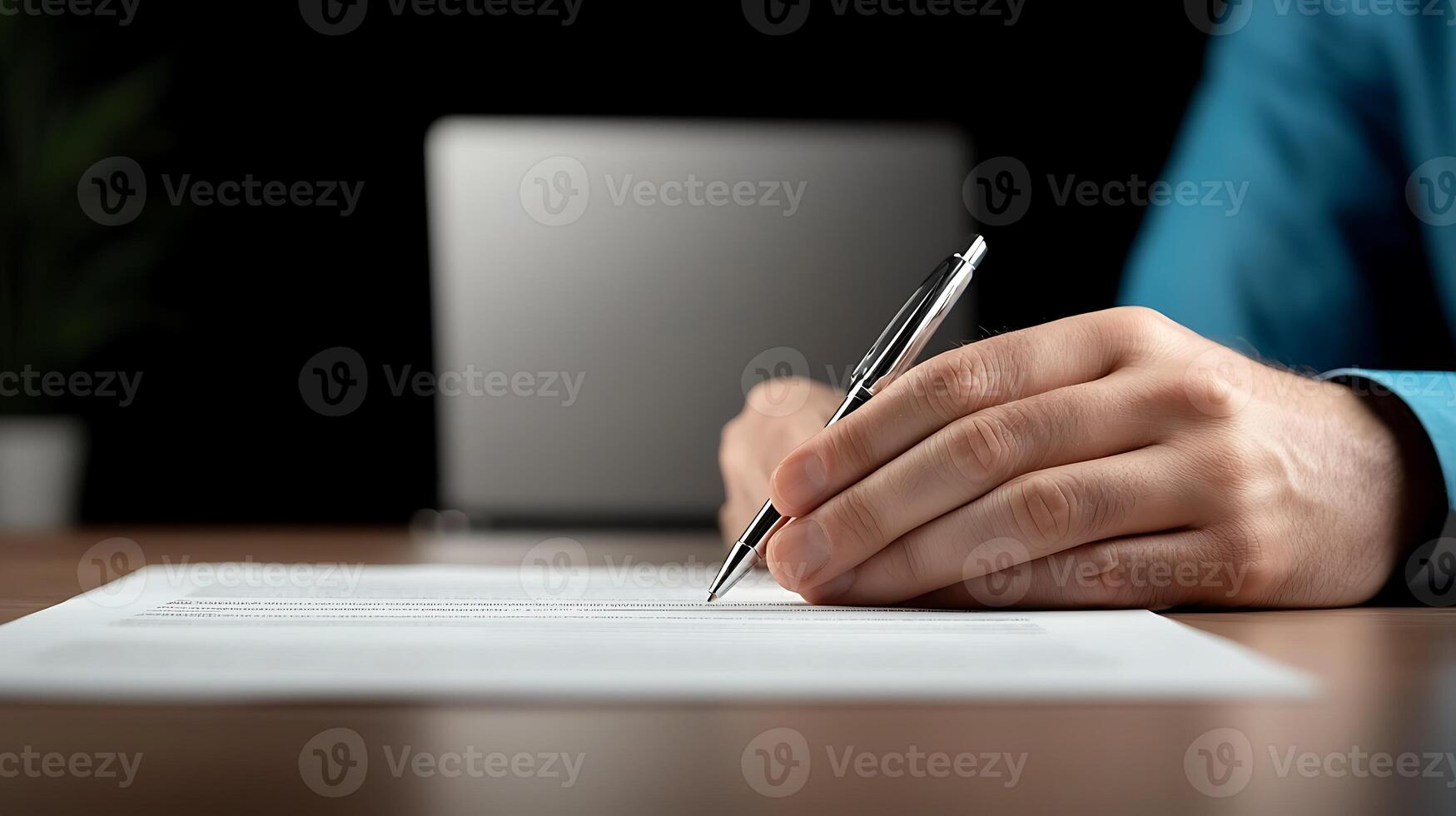 A person in a blue jacket signing a document on a wooden desk with a laptop in the background photo