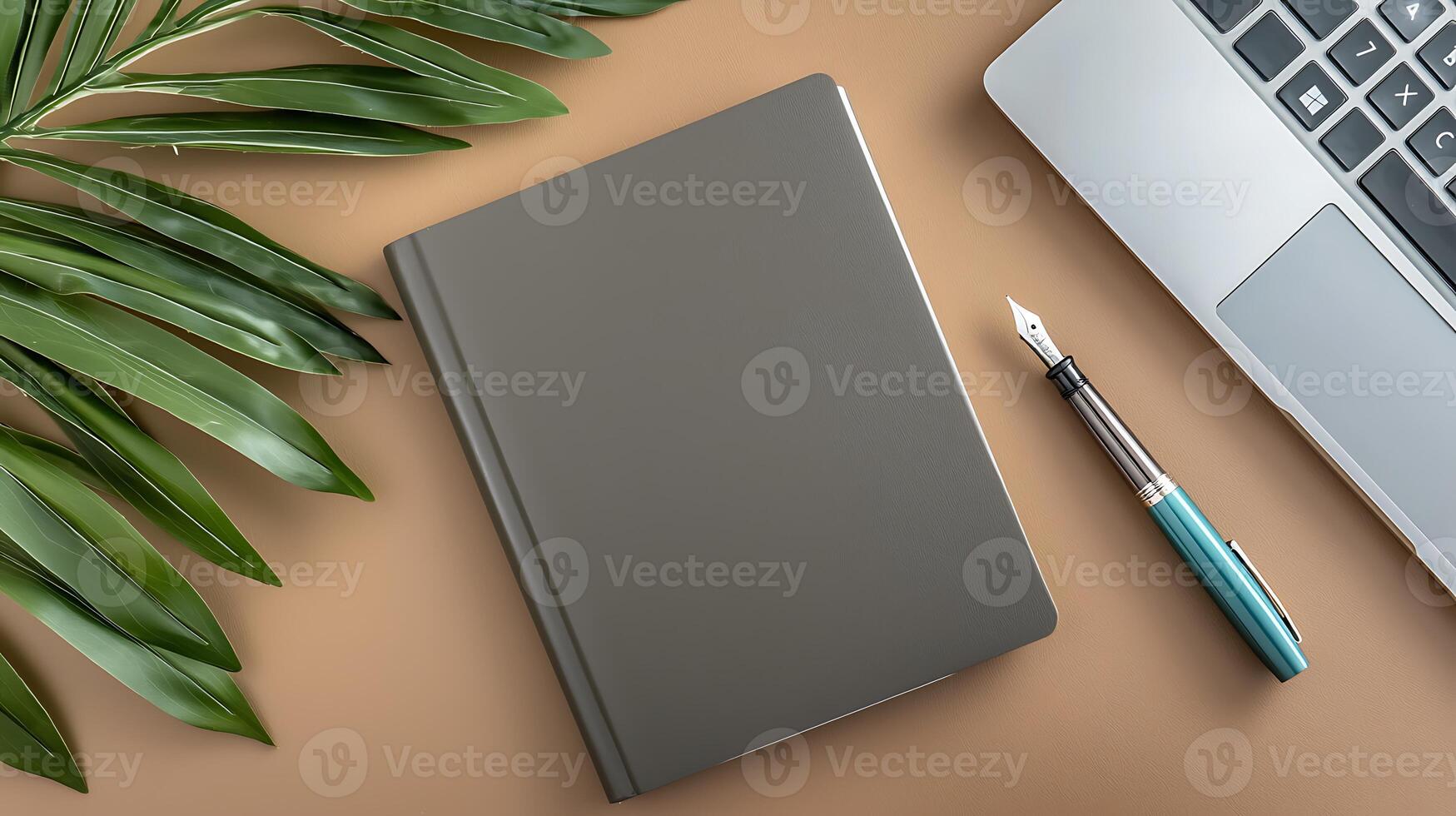 A sleek gray notebook lies on a beige surface next to a laptop and a fountain pen, surrounded by greenery photo