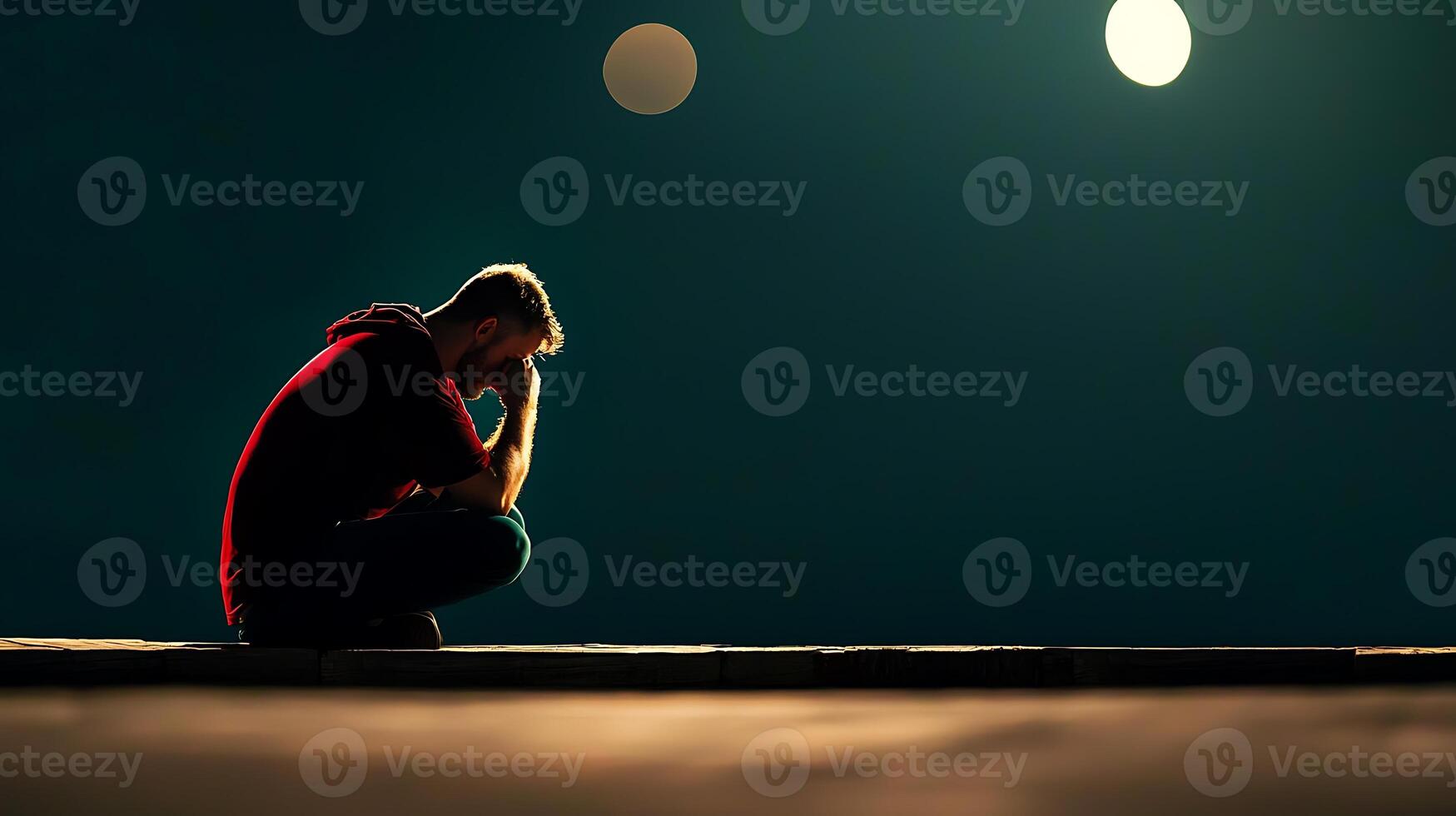 A solitary man in a red hoodie sits on a dark street, deep in thought under dim streetlights photo