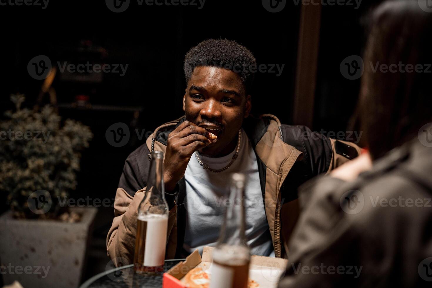 Young man enjoying snack outdoors at night with friends while having drinks and conversing photo