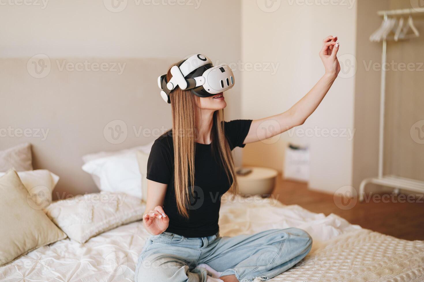 Young woman enjoys virtual reality experience in modern bedroom during daytime while seated on a bed and interacting with surroundings photo