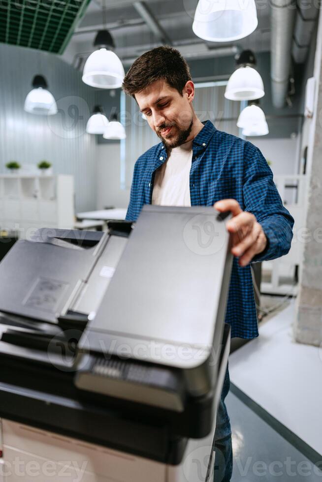 Man operating a copier in a modern office environment during the day while wearing a casual outfit and examining printed documents photo