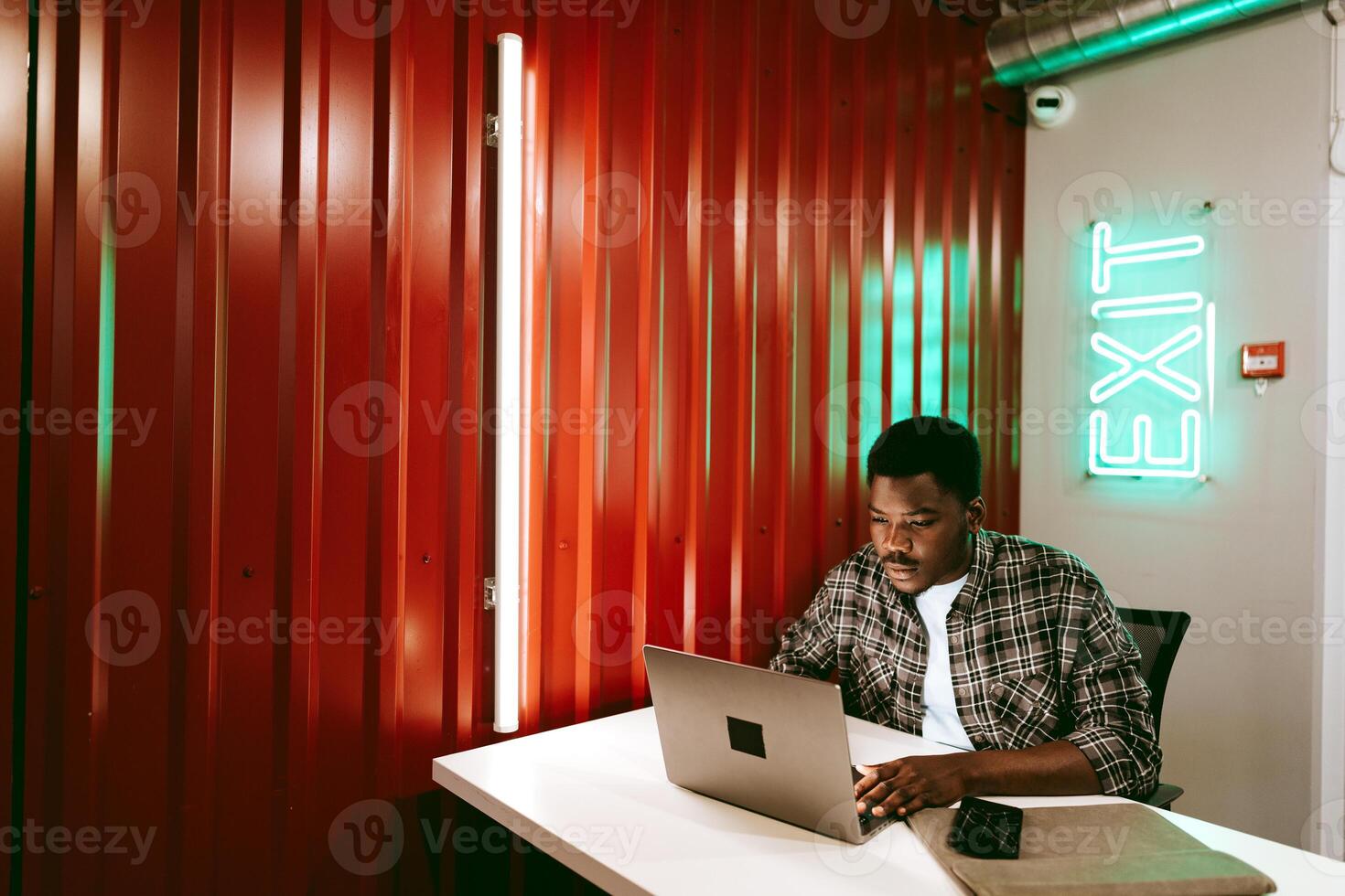 Young man working on a laptop in a modern workspace with colorful neon lights in the background near an exit sign photo