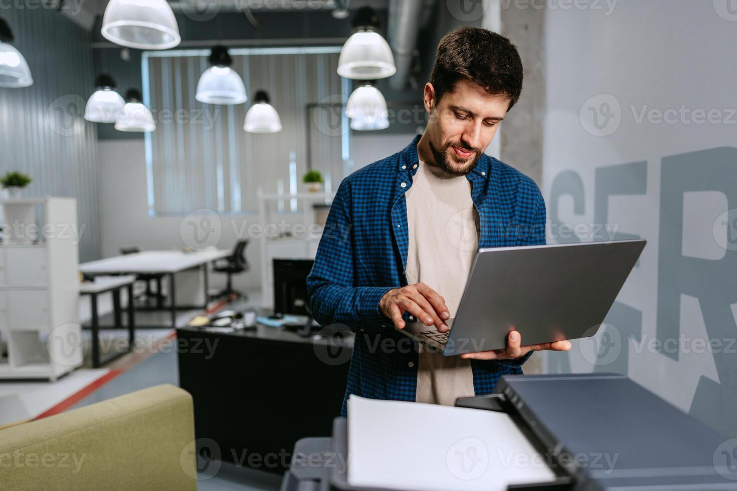 Man working on laptop in modern office setting during daytime while using printer photo