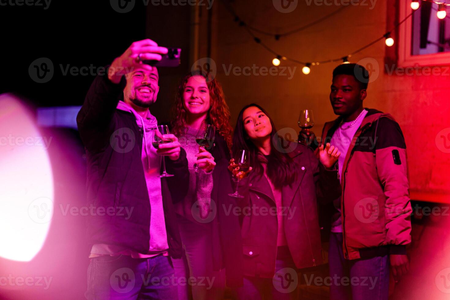 Friends enjoy a lively evening gathering while taking a selfie with drinks in a warmly lit outdoor space photo